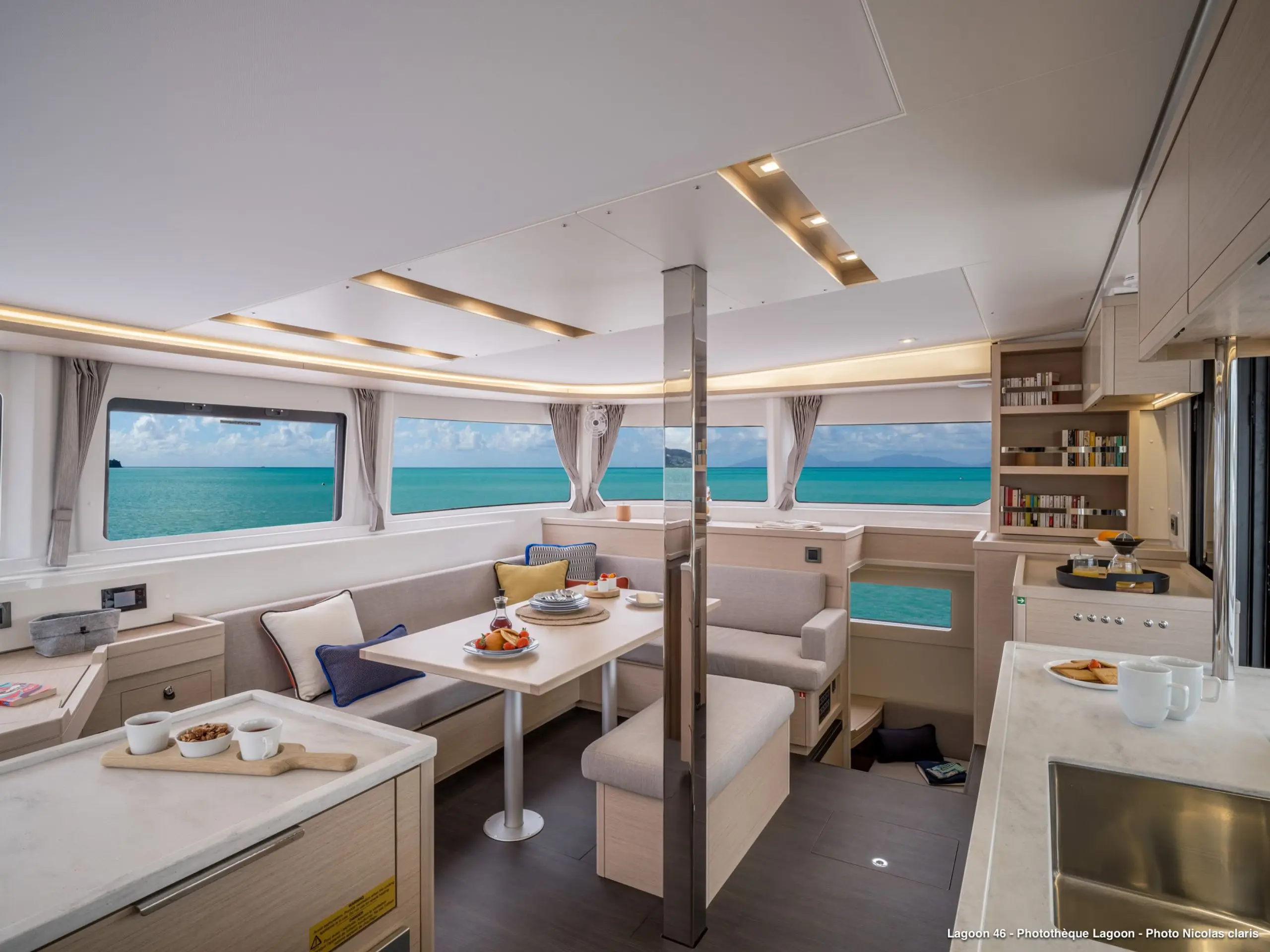 Luxurious Lagoon 46 yacht interior with light wood finishes, a dining table set for breakfast, wraparound windows offering panoramic ocean views, and modern kitchen area on the right. Bright natural light fills the open, cozy space.