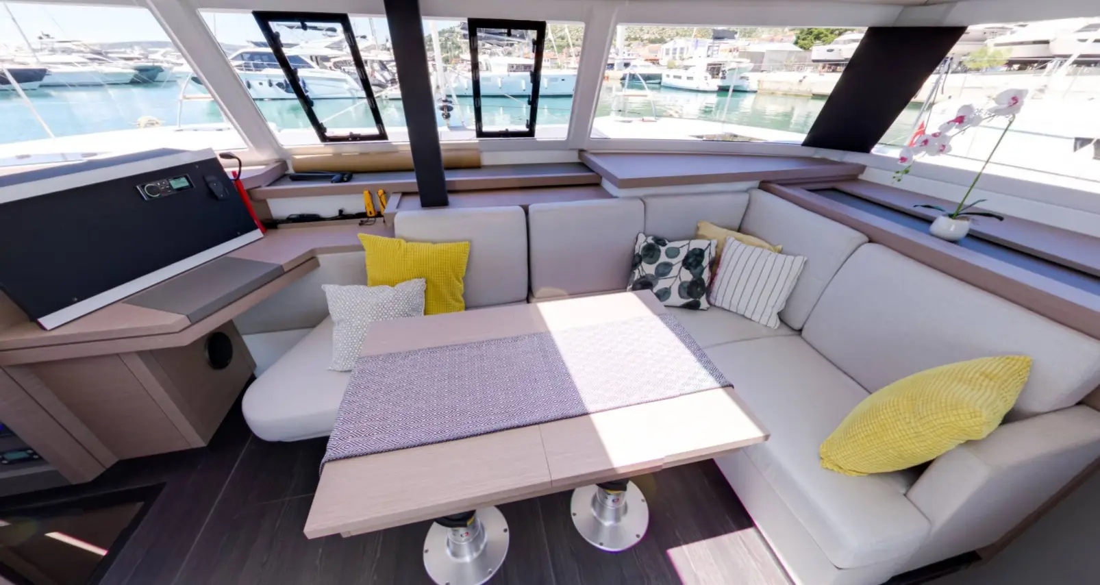 The interior of a Fountaine Pajot Astrea 42 yacht features a U-shaped sofa, colorful throw pillows, a wooden table, large windows with marina views, and a potted orchid. Sunlight brightens the cozy, stylish space.