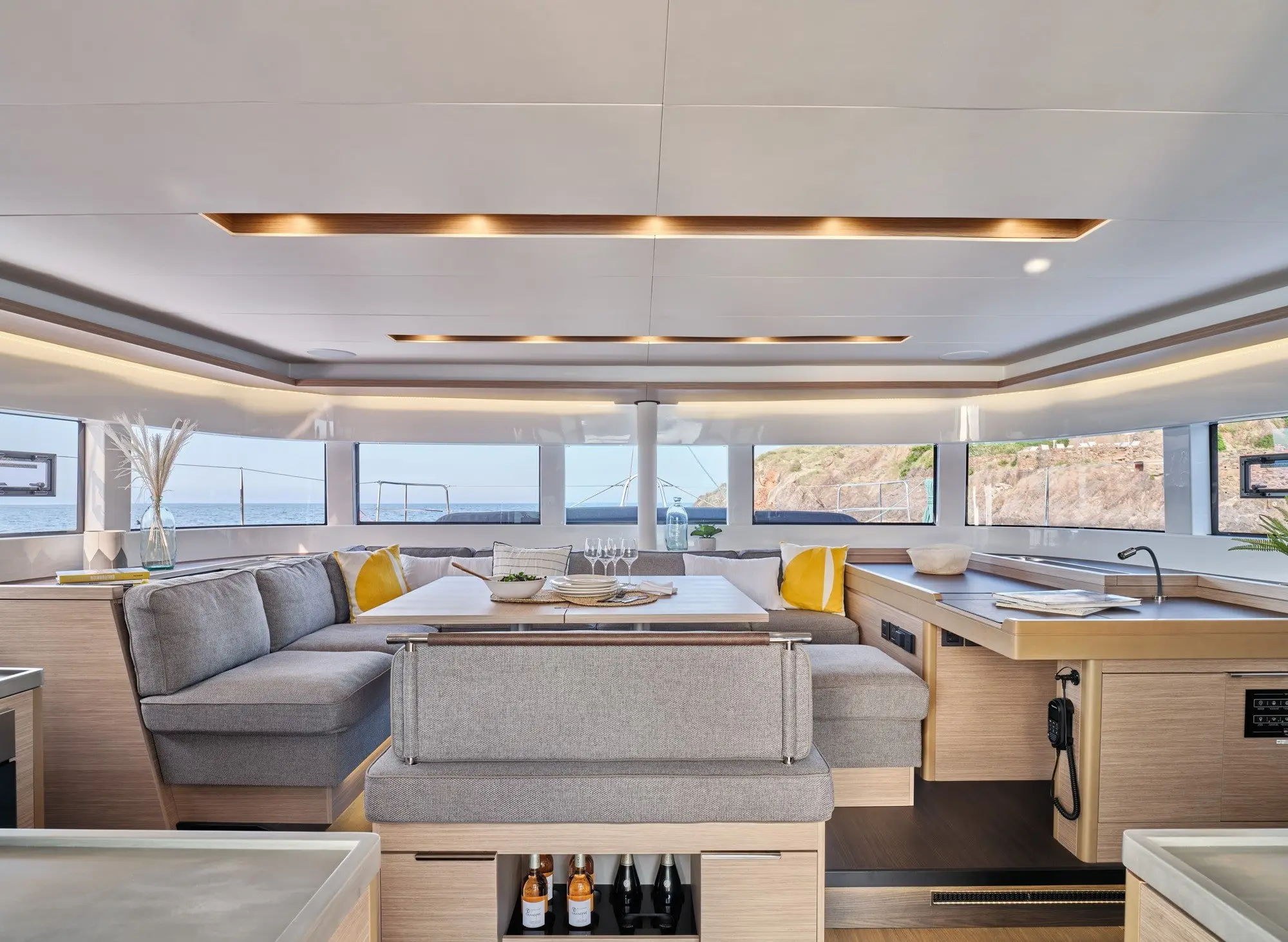 Modern Lagoon 51 yacht interior with large windows, a spacious L-shaped gray sofa, light wood cabinetry, yellow accent pillows, and a table set with glasses, offering views of the sea and coastline outside.