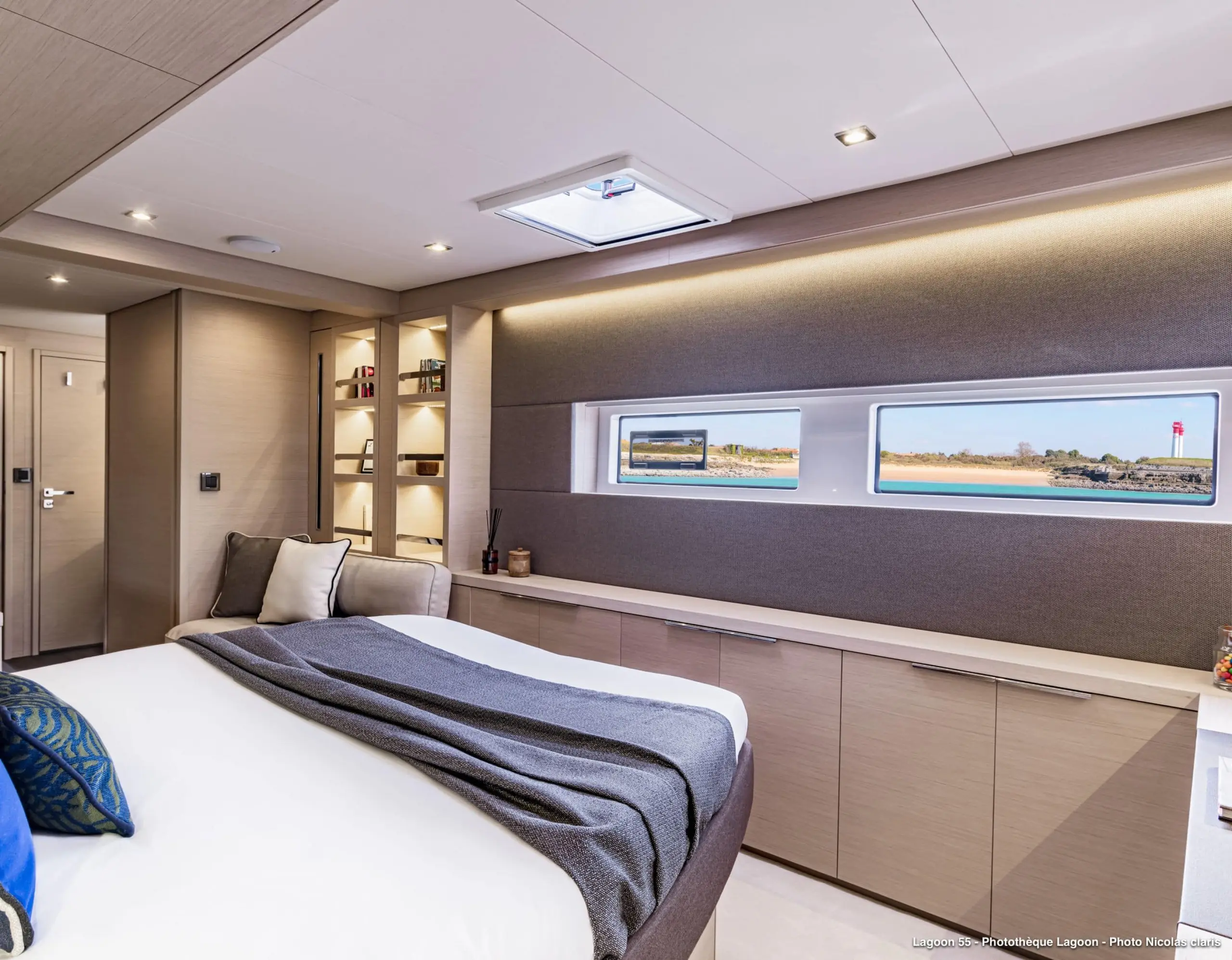 A modern Lagoon 55 yacht bedroom with a large bed, neutral walls, built-in shelves, and rectangular windows revealing a scenic water view. Soft lighting and minimalistic decor create a cozy, luxurious atmosphere.