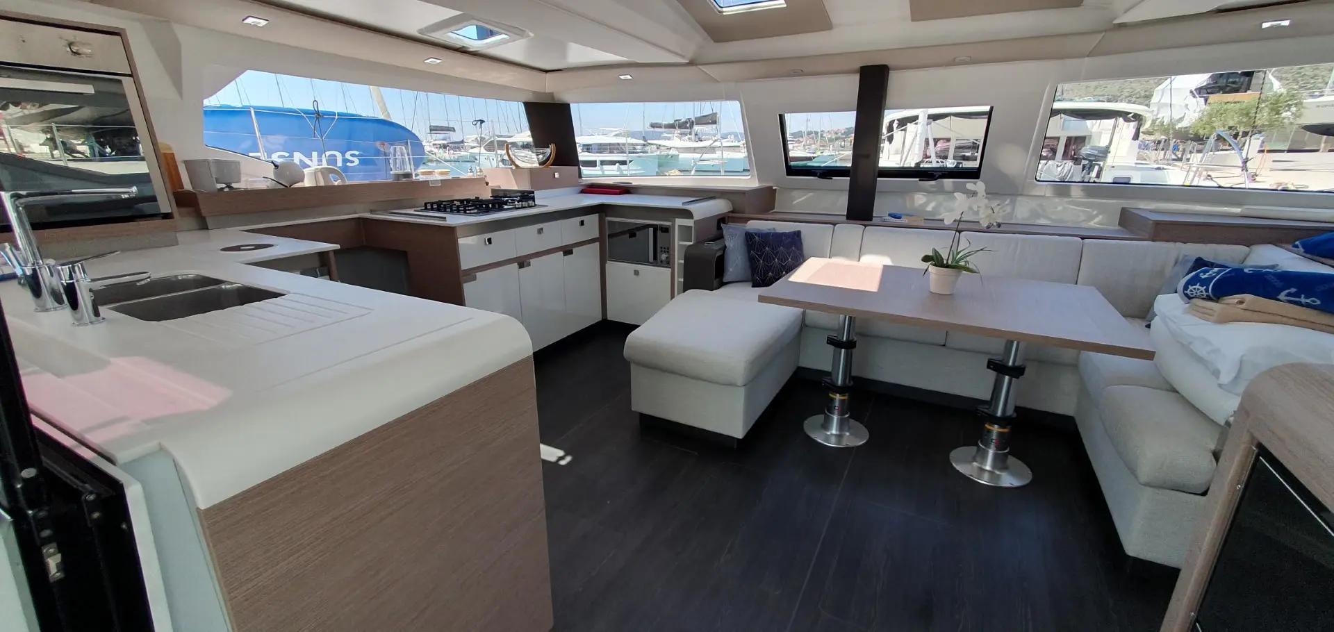 The interior of a modern Fountaine Pajot Elba 45 yacht features a kitchen with a stove and sink, plus a dining area with a table, cushioned benches, and windows overlooking a marina filled with docked boats.