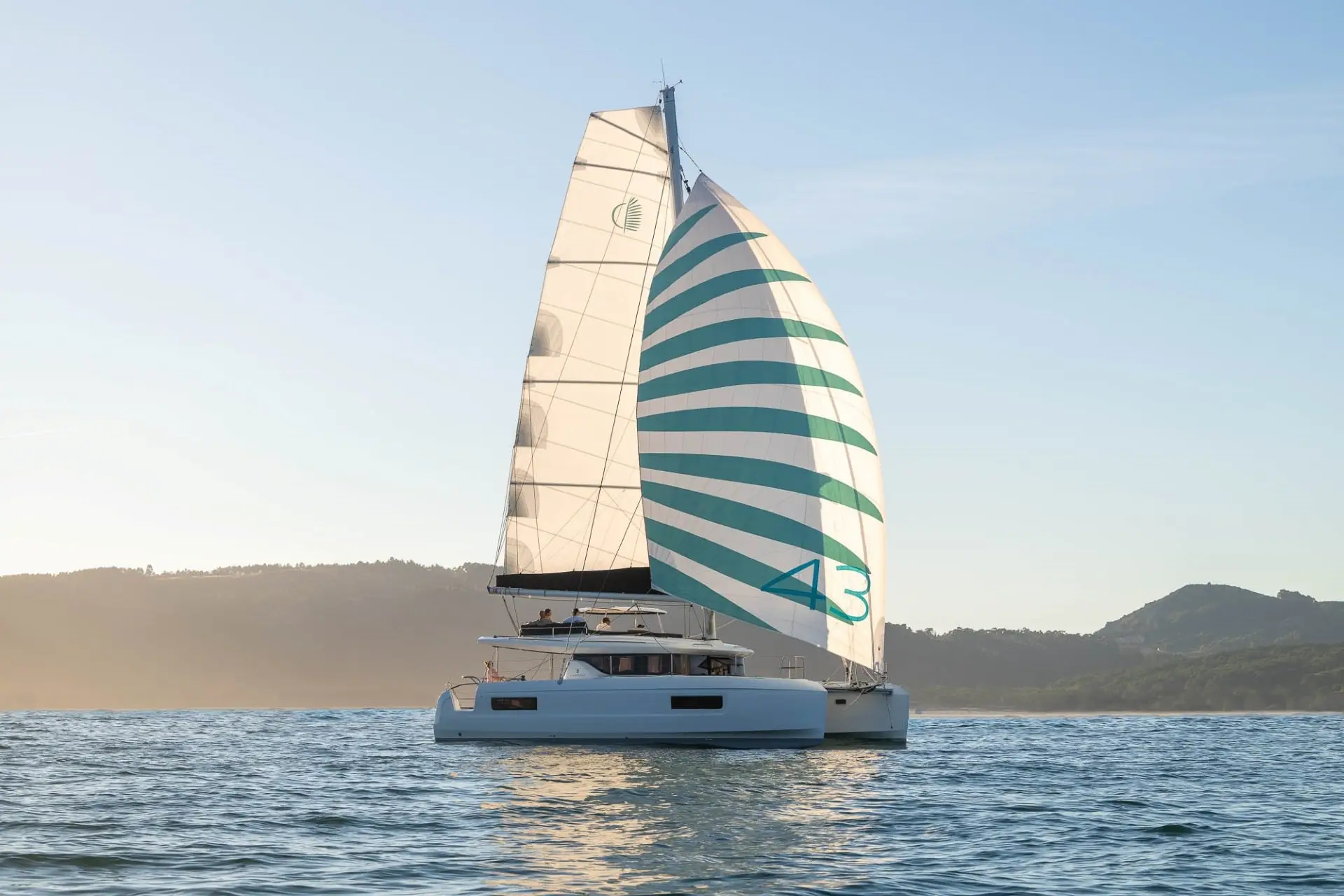 A white Lagoon 43 catamaran with a large, striped sail featuring the number 43 glides on calm blue water. Hills and a hazy sky are visible in the background under soft sunlight.