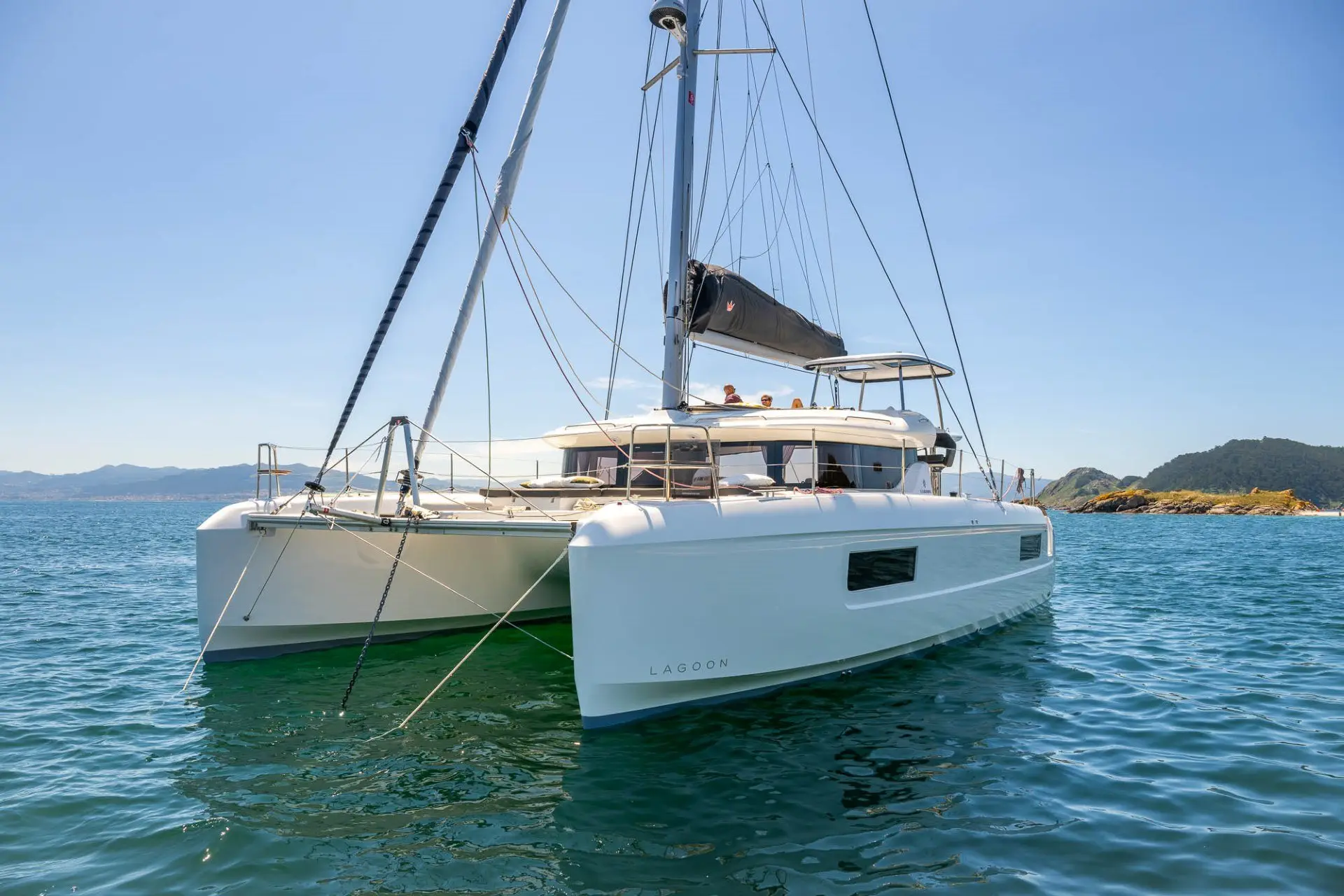 A modern white Lagoon 43 catamaran yacht floats on calm blue water near a rocky coastline under a clear sky, with a few people visible onboard enjoying the sunny day.