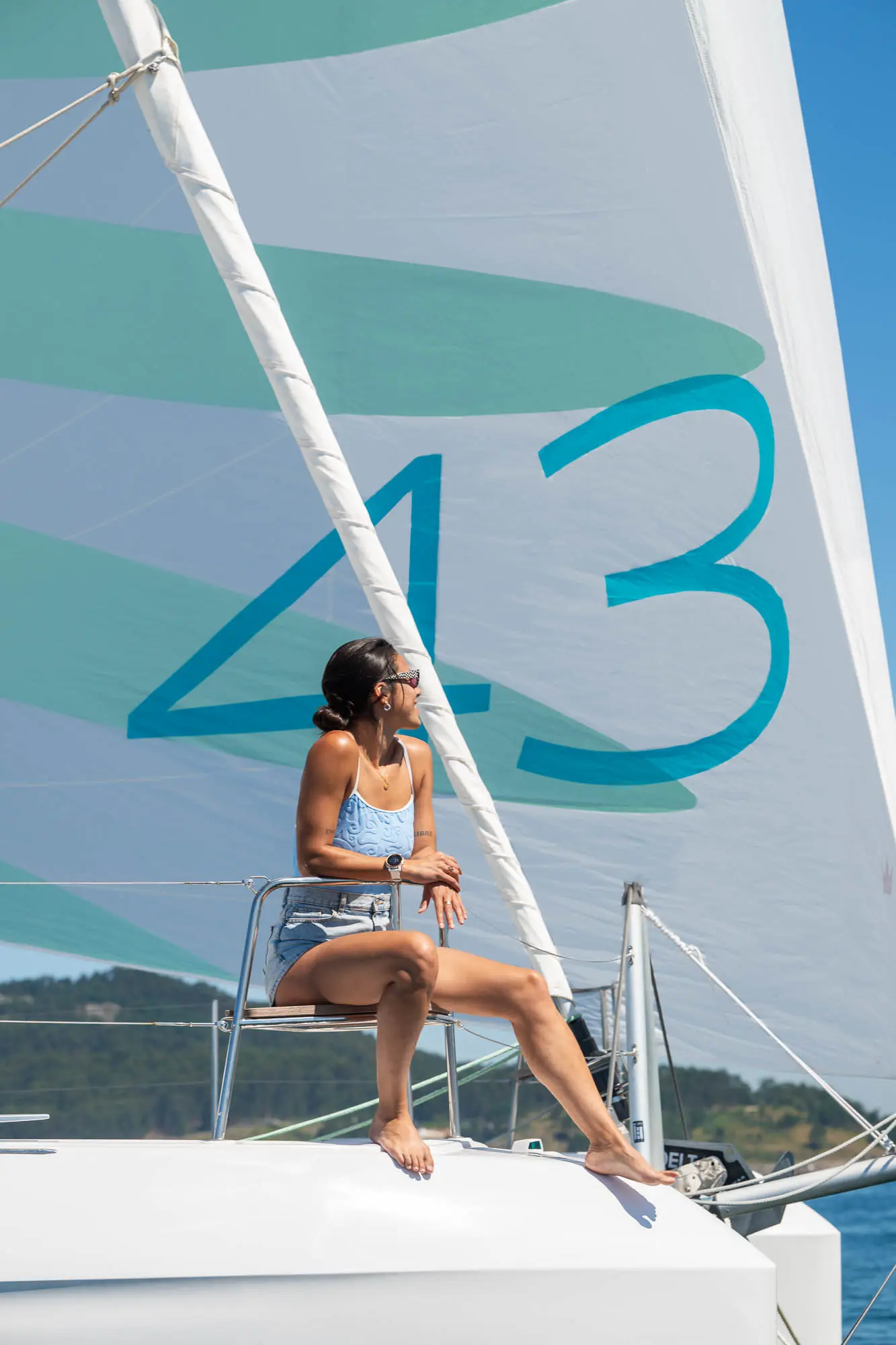 A woman in a swimsuit sits relaxed on a Lagoon 43 sailboat, looking to the side. The large sail behind her has the number 43 printed on it, with hills and greenery visible in the distant background.