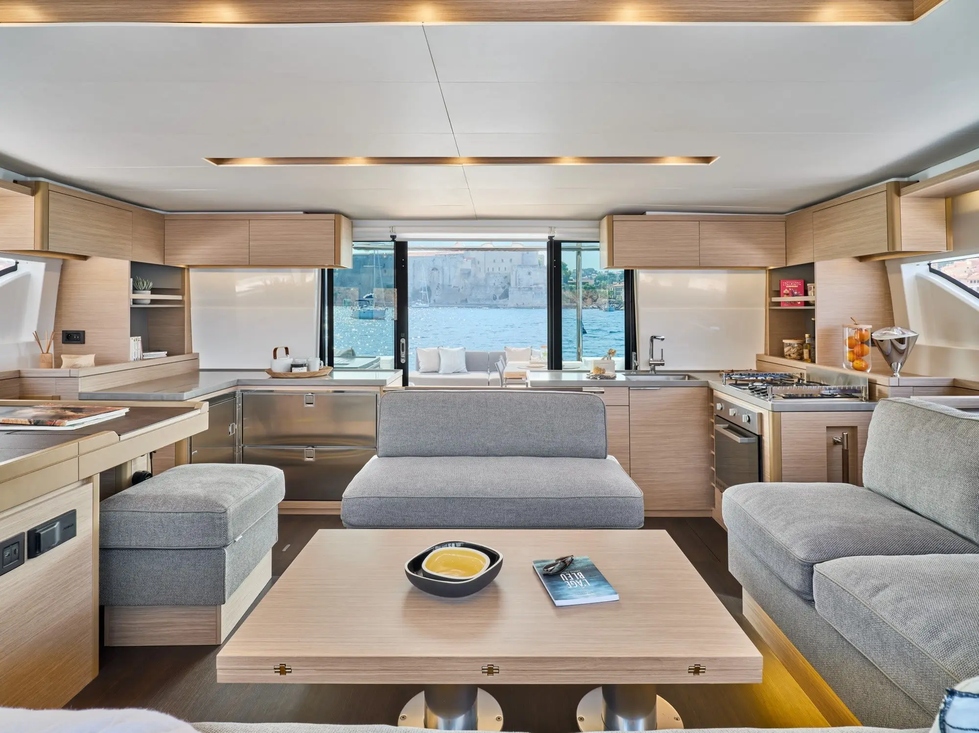 Modern Lagoon 51 yacht interior with light wood cabinetry, grey upholstered seating, and a central coffee table with a bowl and magazine. Large windows offer water views and a distant building outside. The space is bright and stylish.