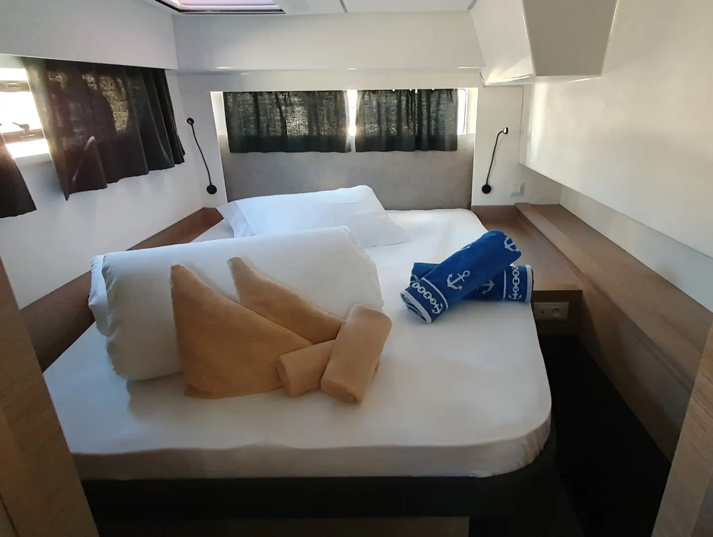 A neatly made bed in a small, modern room with pillows, folded tan towels, and two blue towels with anchor designs aboard the Fountaine Pajot Elba 45. The room features dark curtains, white walls, and wood accents.