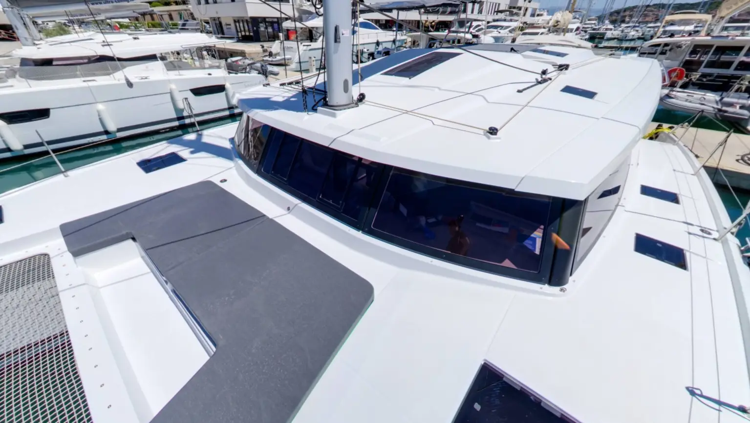 Close-up view of the deck and cockpit windows of a white Fountaine Pajot Astrea 42 catamaran docked at a marina, with other boats and buildings visible in the background.