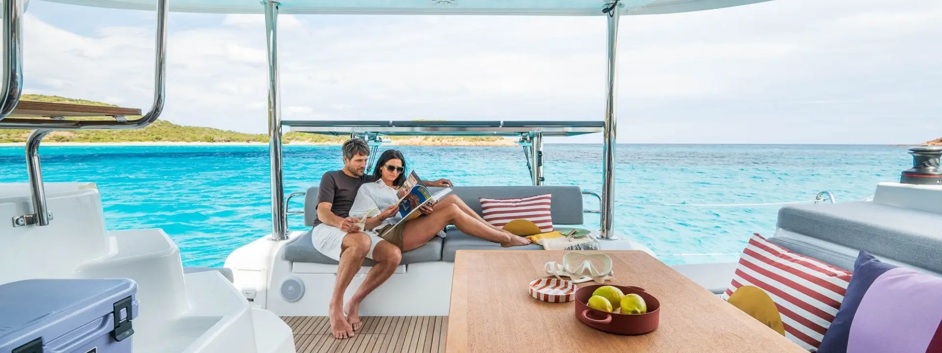A couple relaxes on a Lagoon 38 yacht, lounging on cushioned seats with striped pillows, reading a magazine. Clear turquoise water and a distant shoreline are visible in the background. A bowl of fruit sits on the wooden table nearby.