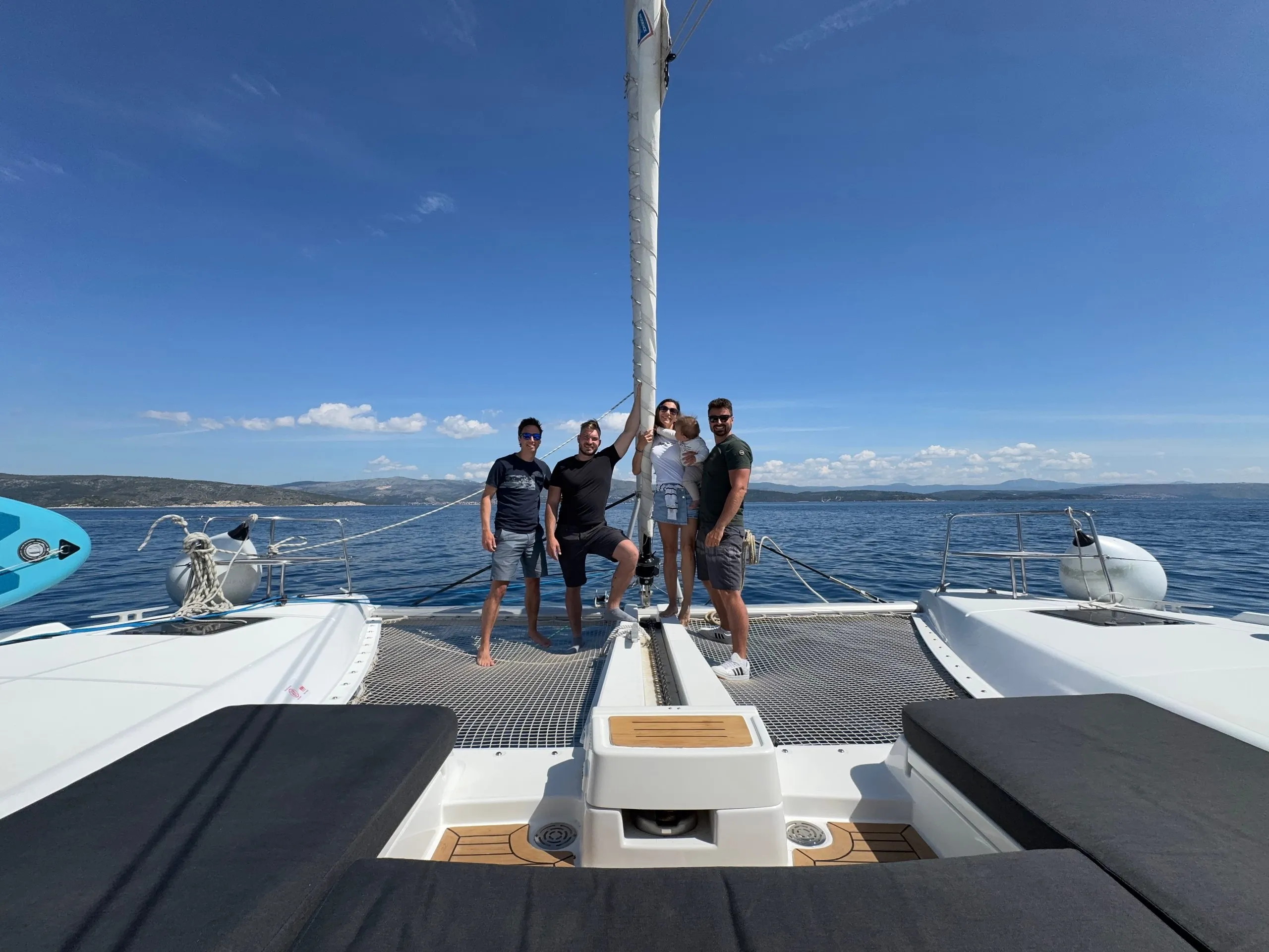 Four people stand smiling on the deck of a Lagoon 46 catamaran sailboat under a clear blue sky, with calm sea and distant coastline in the background.