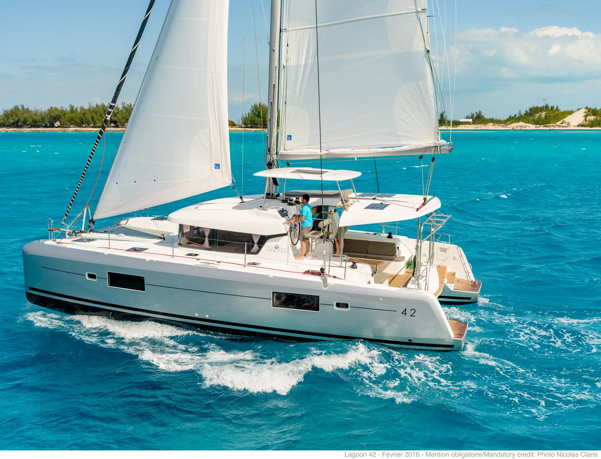 A white catamaran sailboat with two people on deck glides over clear turquoise water near a sandy, tree-lined shore under a sunny blue sky.