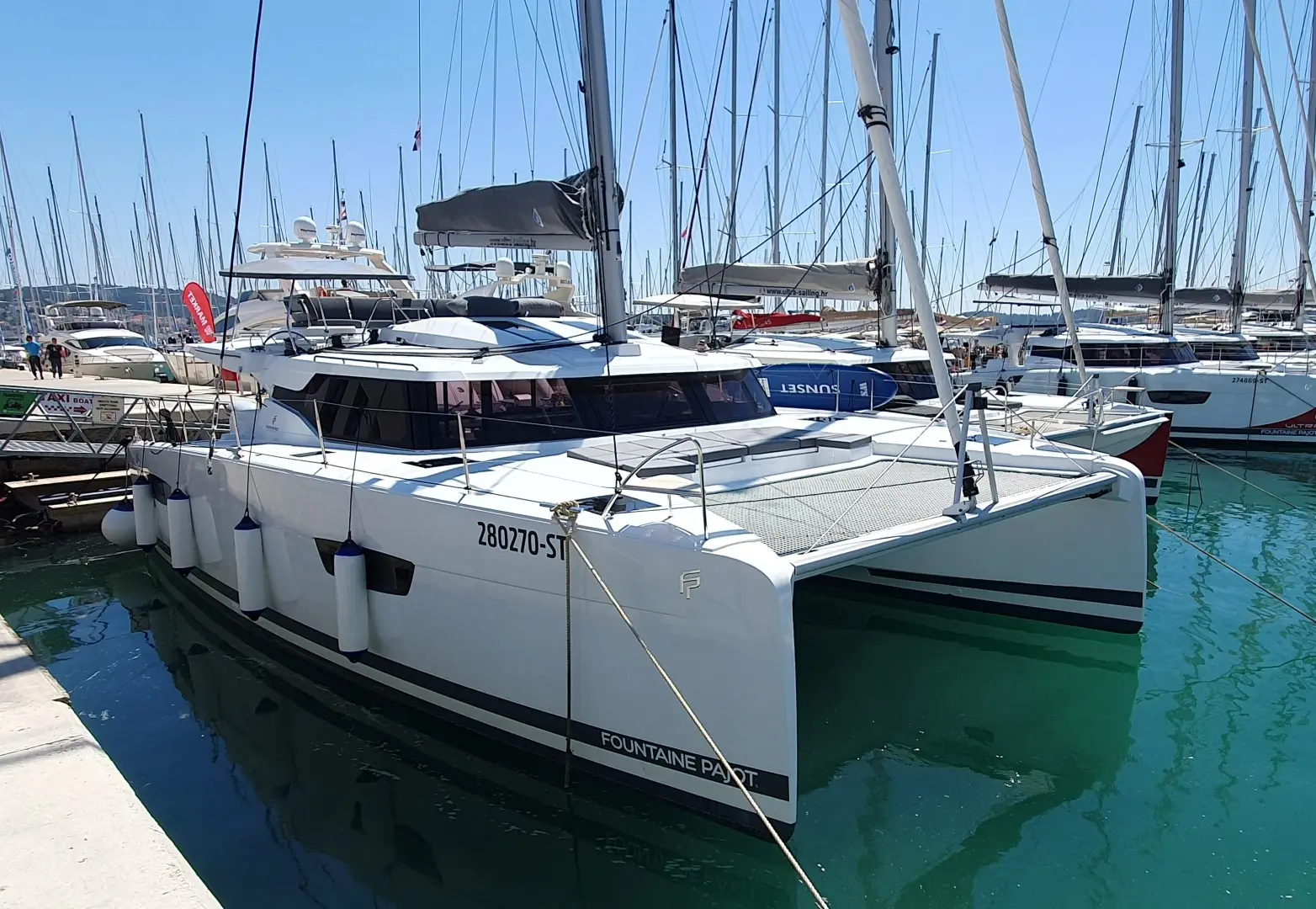 A modern white catamaran, the Fountaine Pajot Elba 45, is docked in a marina, surrounded by other sailboats on a sunny day with clear blue skies and calm greenish water.