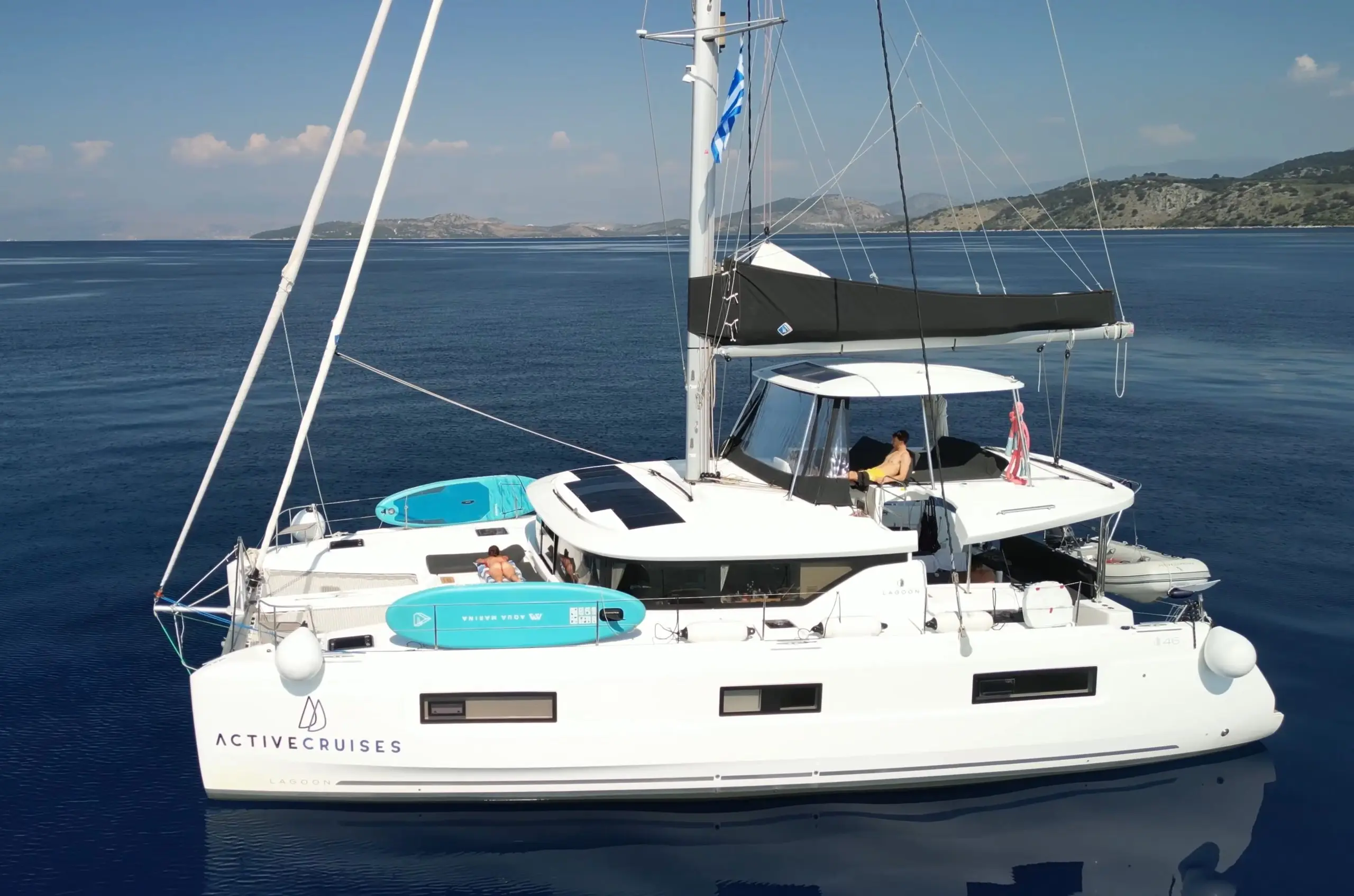 A white Lagoon 46 sailing catamaran with Active Cruises branding floats on calm blue water. Two paddleboards are secured on deck and a person relaxes under the shaded cockpit. Distant islands are visible under a clear sky.
