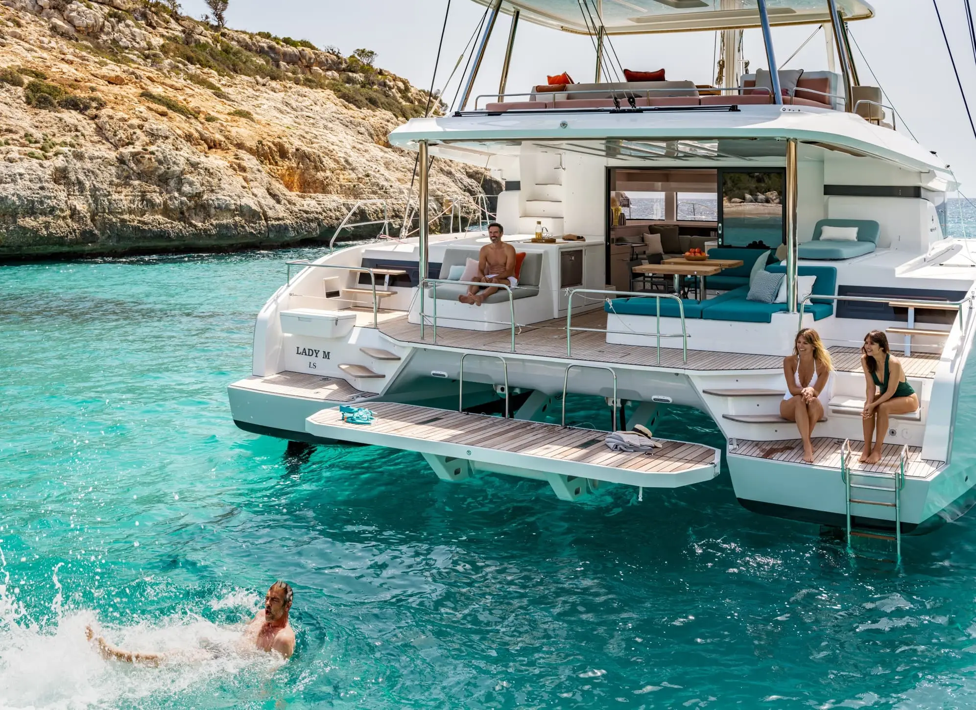 A luxurious Lagoon 55 white catamaran anchored in clear turquoise water, with people relaxing on deck and one person swimming nearby; rocky coastline and blue sky are visible in the background.