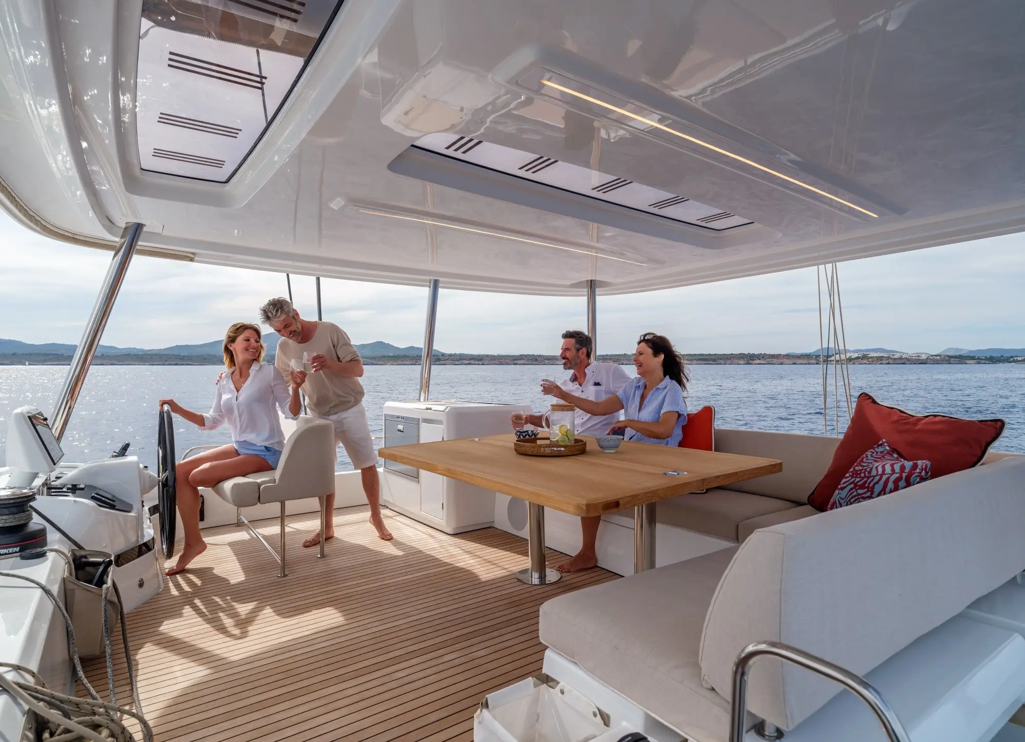 Four people relax on a Lagoon 55’s covered deck. Two are seated at a table with drinks and snacks, while another couple stands near the helm. The sea and distant hills are visible in the background.