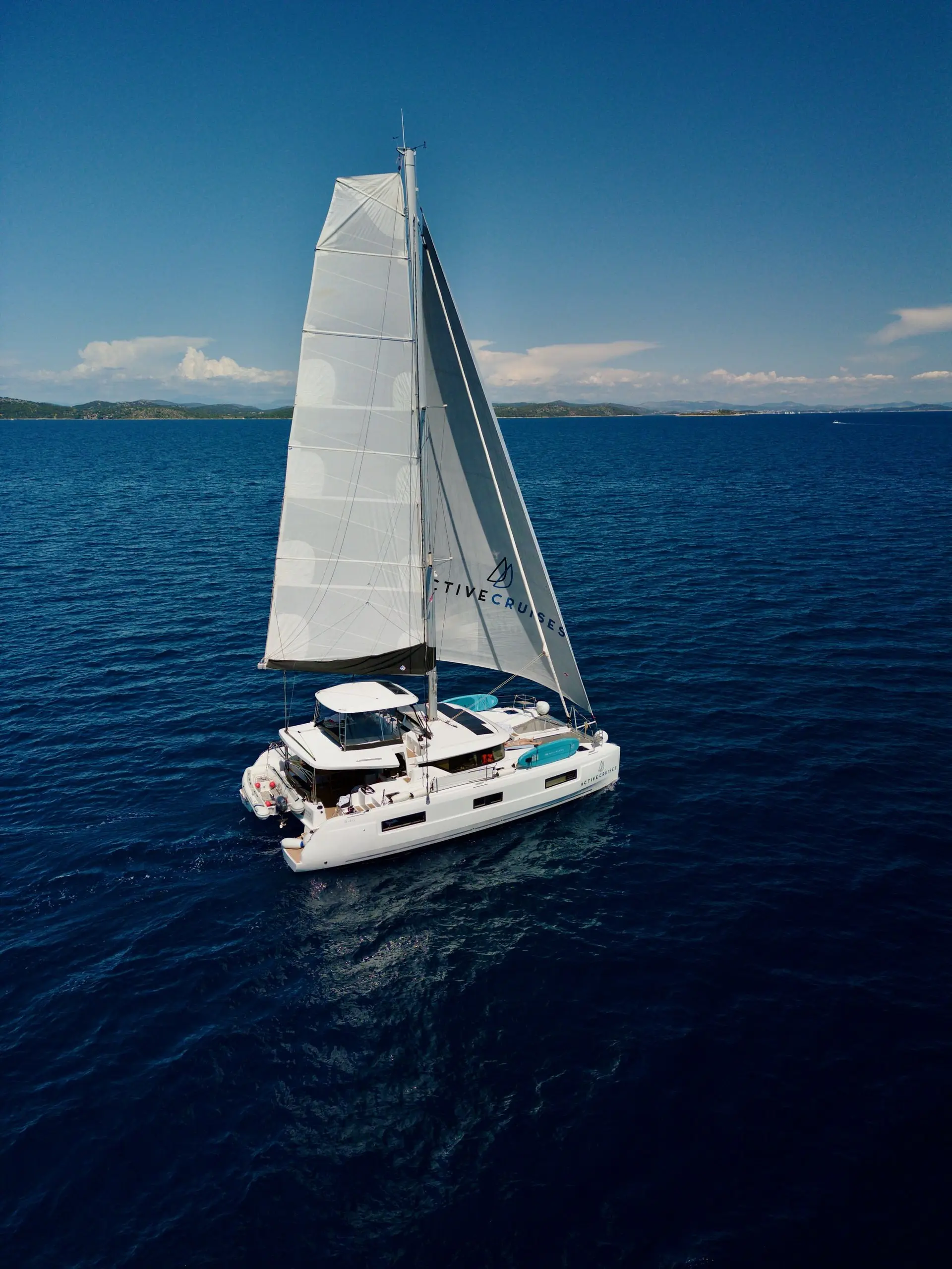 A white Lagoon 46 catamaran sailboat with tall sails glides across deep blue ocean water on a sunny day, with distant land and a clear sky in the background.