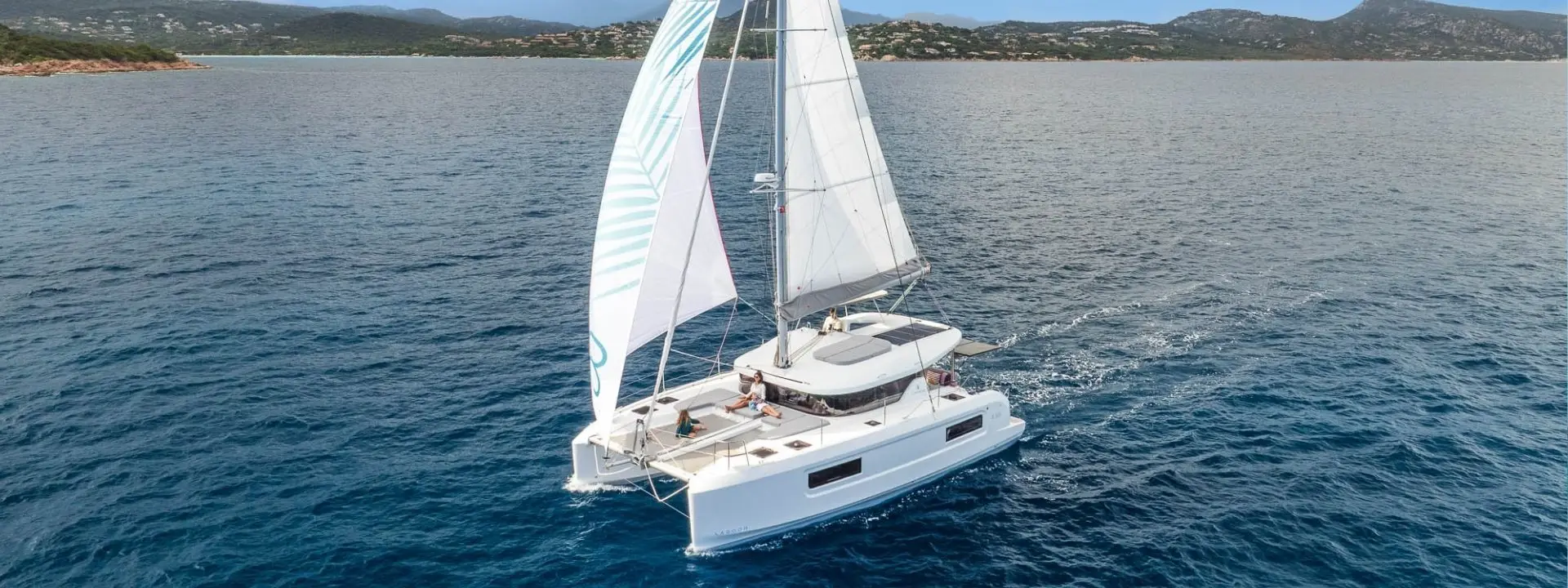 A white Lagoon 38 catamaran sailboat with raised sails glides on calm blue water near a mountainous coastline with scattered greenery under a clear sky.
