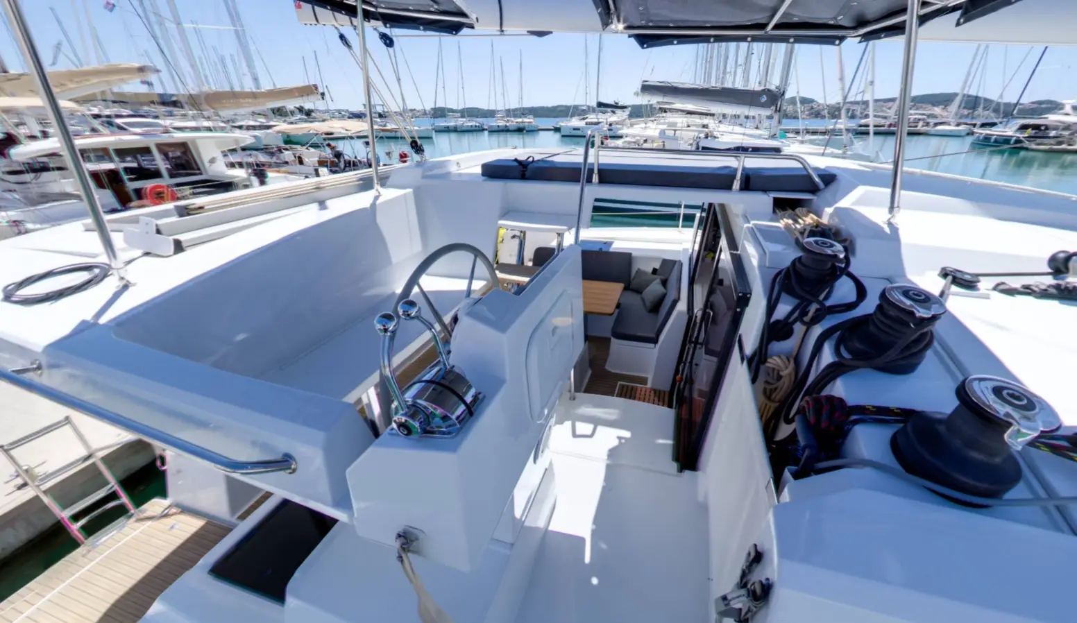 View of a Fountaine Pajot Astrea 42 yacht’s cockpit with a steering wheel, control panels, winches, ropes, shaded seating area, and docked boats and masts visible in the marina background under a clear sky.