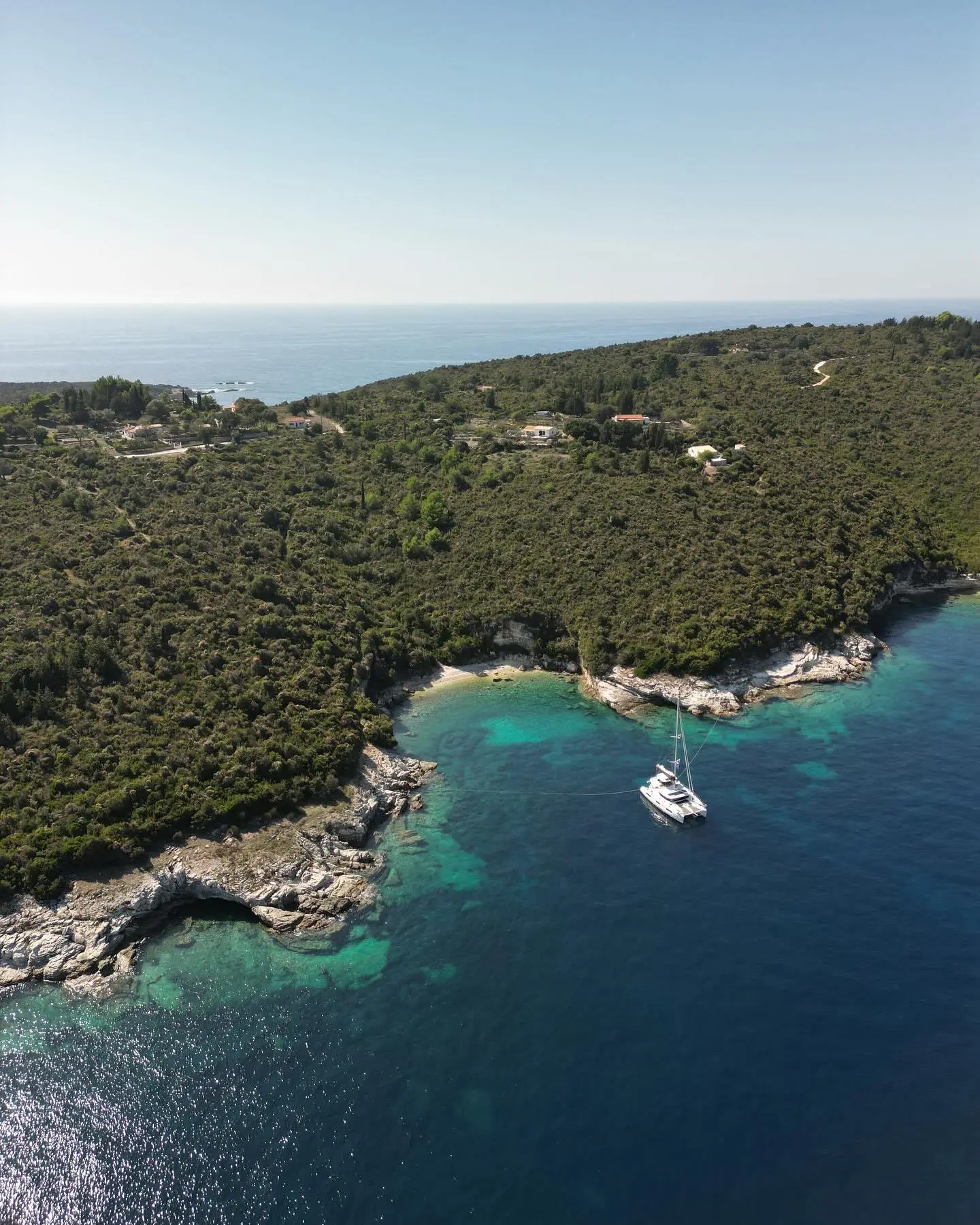 Aerial view of a Lagoon 43 catamaran anchored in turquoise water near a green, rocky coastline.