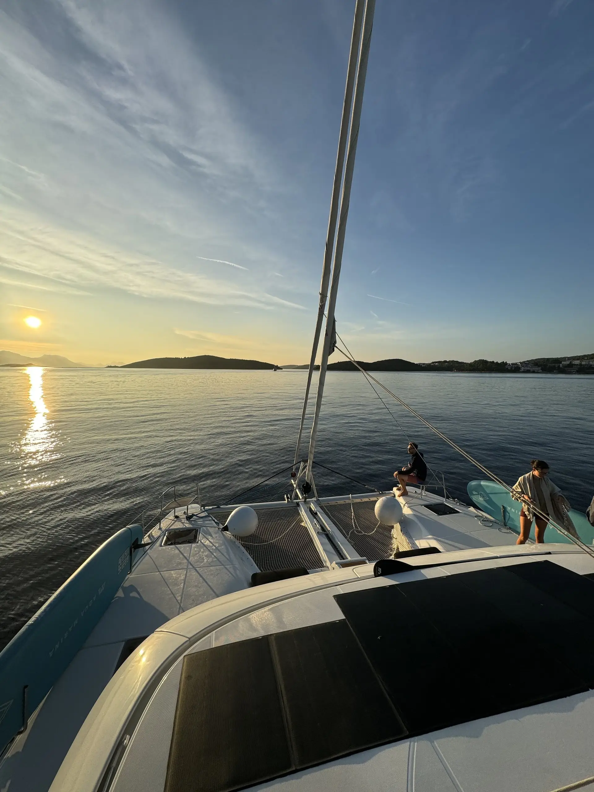 View from the deck of a Lagoon 46 catamaran sailing on calm water at sunset, with two people near the bow. The sky is clear, and distant islands reflect golden sunlight on the sea.