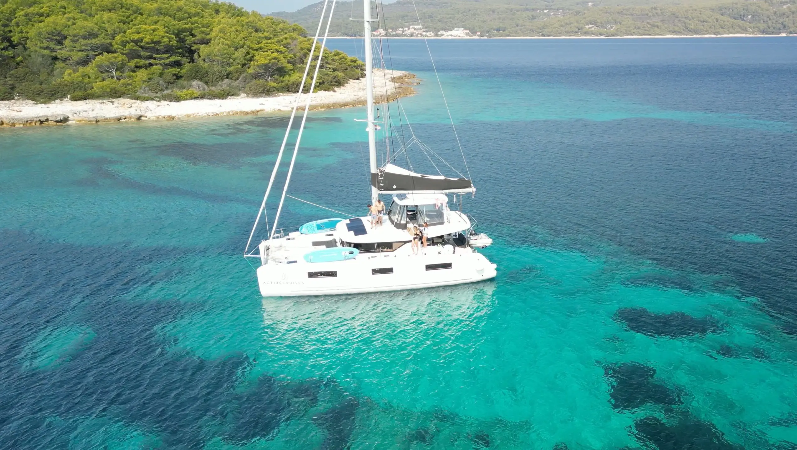 A white Lagoon 46 catamaran yacht floats on clear, turquoise water near a rocky, tree-lined shoreline with lush greenery under a bright sky. The water shows varying shades of blue and reveals the shallow seabed.