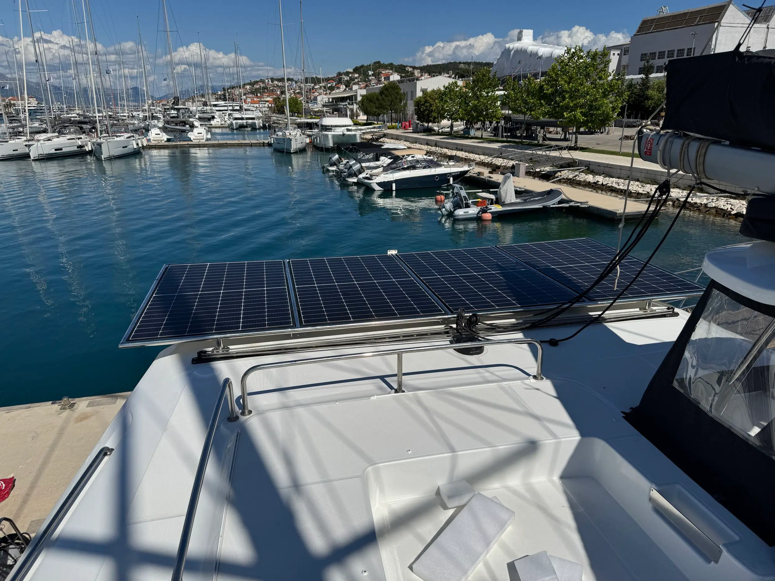 View from a Lagoon 43 docked at a marina, featuring solar panels on the boat’s roof, calm water, several docked boats, and a coastal town with hills and buildings under a clear blue sky.