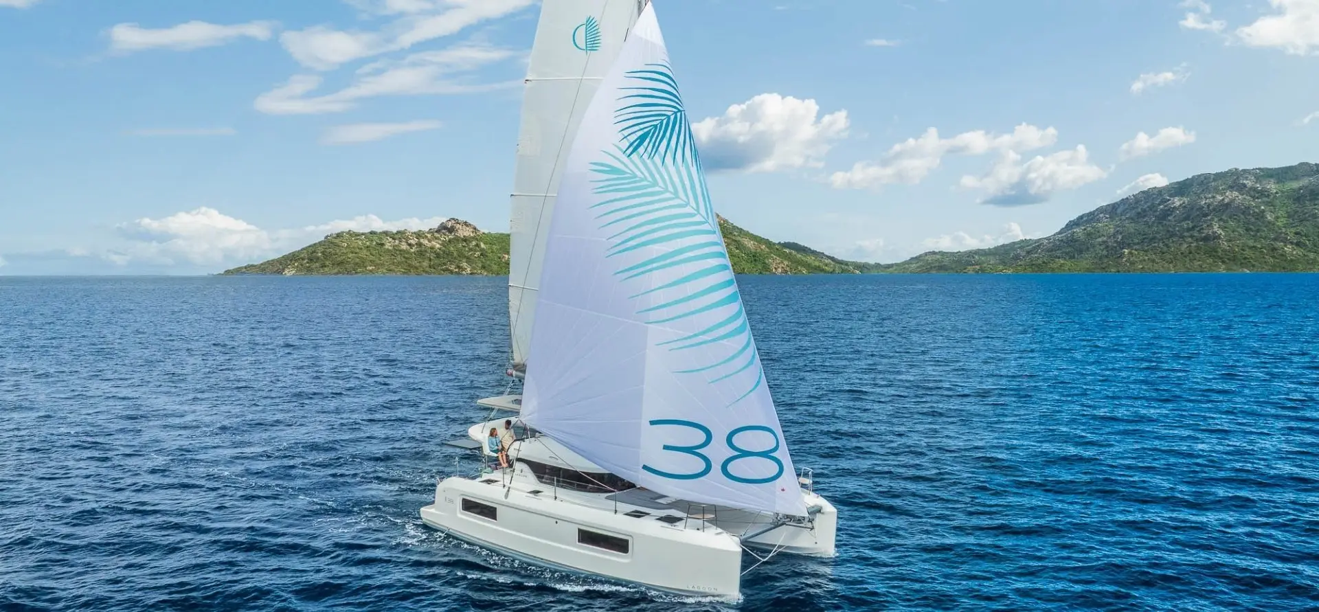 A white Lagoon 38 sailing catamaran with the number 38 on its sail glides across deep blue water, with green, hilly islands in the background under a partly cloudy sky.