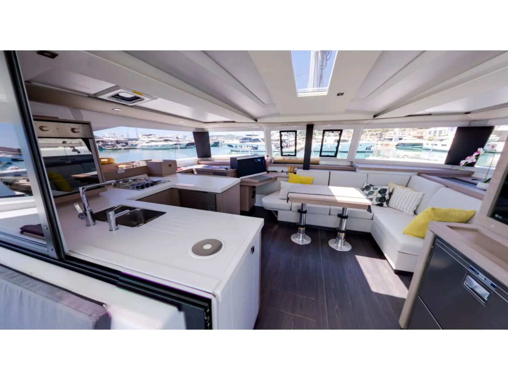 Modern Fountaine Pajot Astrea 42 yacht interior with large windows, a kitchen area with a sink and stove, and a spacious seating area with white couches, yellow pillows, and dining table overlooking the marina.