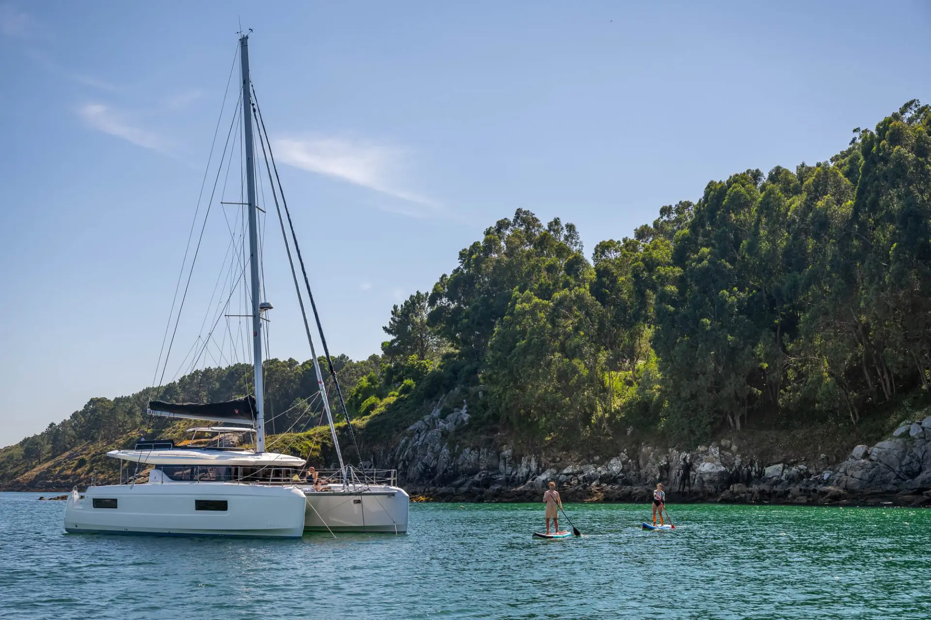 A white Lagoon 43 sailboat is anchored near a forested shoreline, while two people paddleboard on calm turquoise water under a clear blue sky.