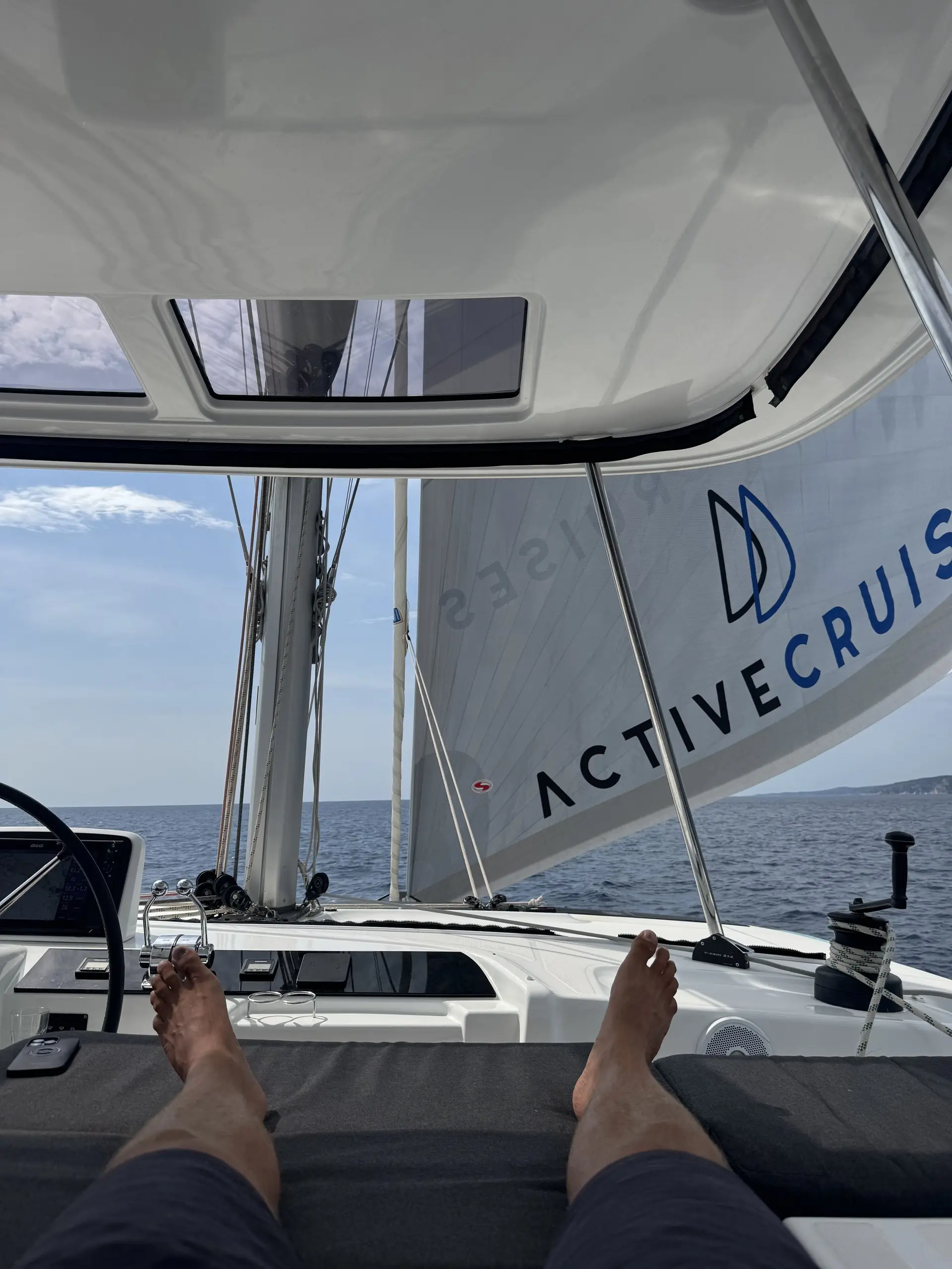 A person relaxes on a Lagoon 46 sailboat, feet up, with a view of the ocean and a large sail reading “ACTIVE CRUISE” on a sunny day. The scene is peaceful, perfectly capturing sailing and leisure.