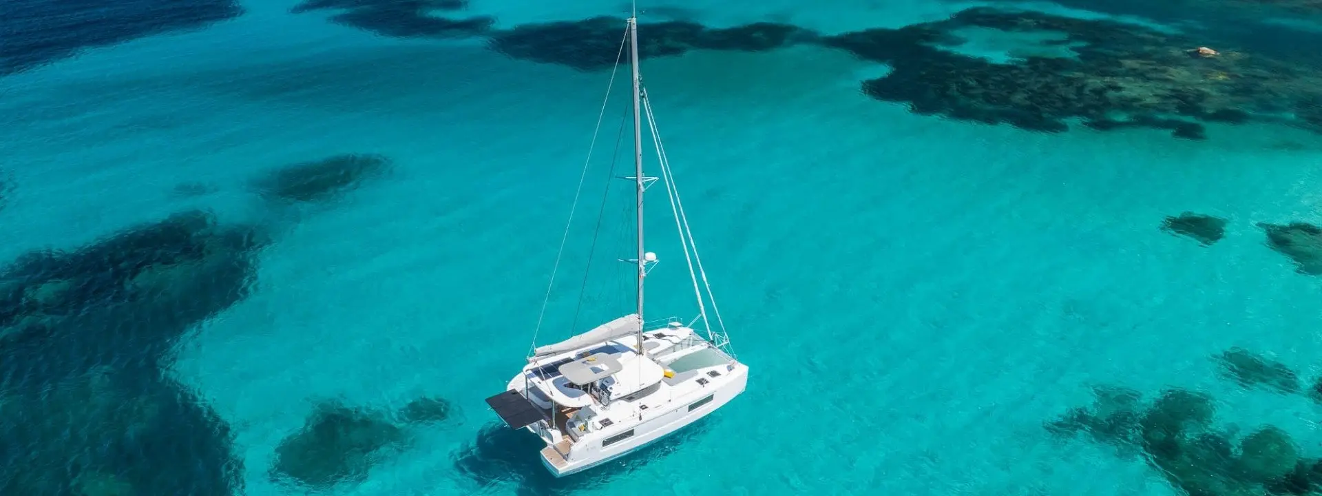 A white Lagoon 38 catamaran sailboat floats on clear turquoise waters, surrounded by patches of dark coral reefs visible beneath the surface, under bright sunlight.