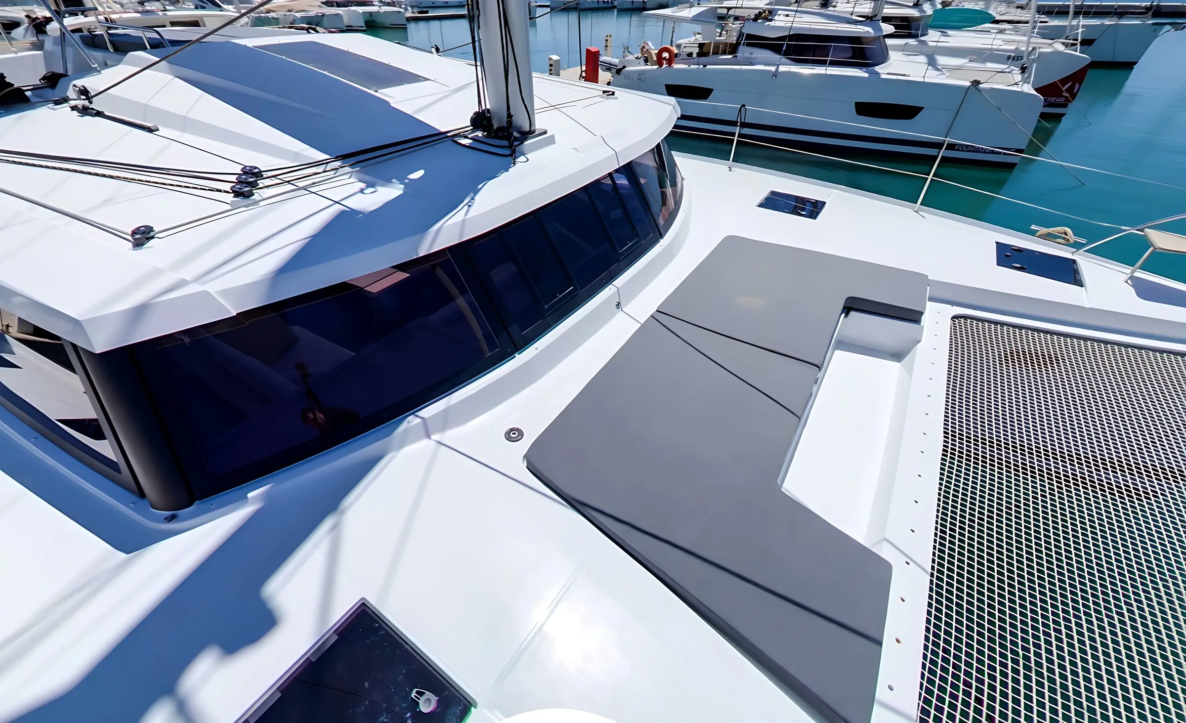 A close-up view of a modern white Fountaine Pajot Astrea 42 catamaran yacht docked at a marina, featuring a shaded cabin, cushioned seating area, and netted trampoline with other yachts moored in the background.