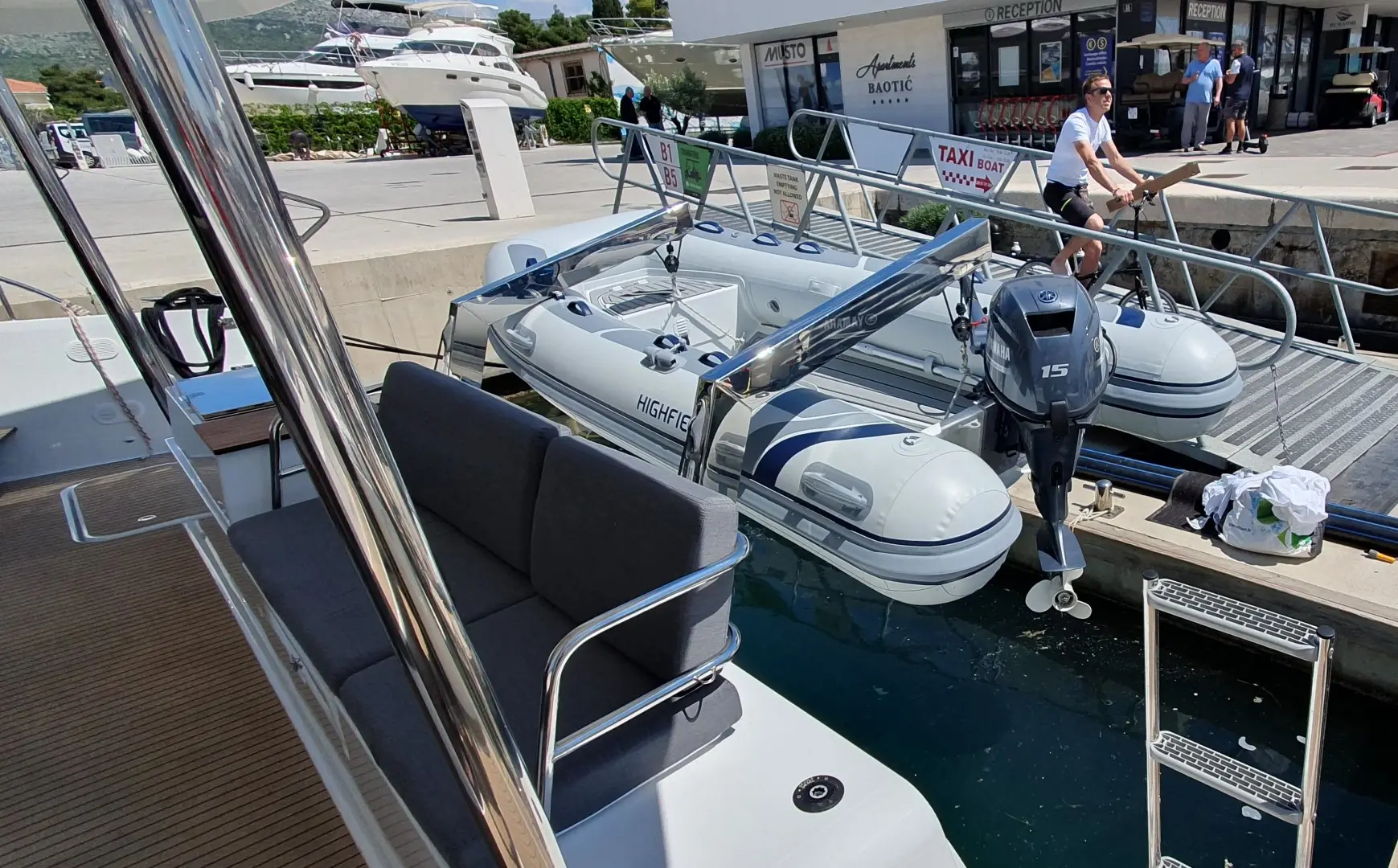 A small inflatable boat with an outboard motor is docked at a marina near a Fountaine Pajot Elba 45. A woman stands on the pier, with benches on a larger boat in the foreground and yachts in the background.