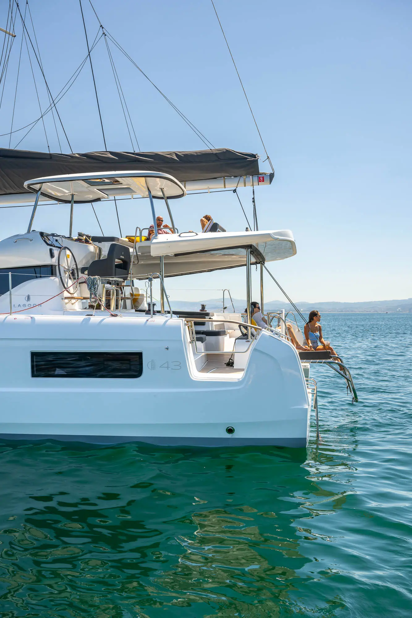 A white Lagoon 43 catamaran yacht floats on calm blue water. Two people relax on the rear steps, dipping their feet in the sea, while others sit under the shade canopy on the upper deck. The sky is clear and sunny.