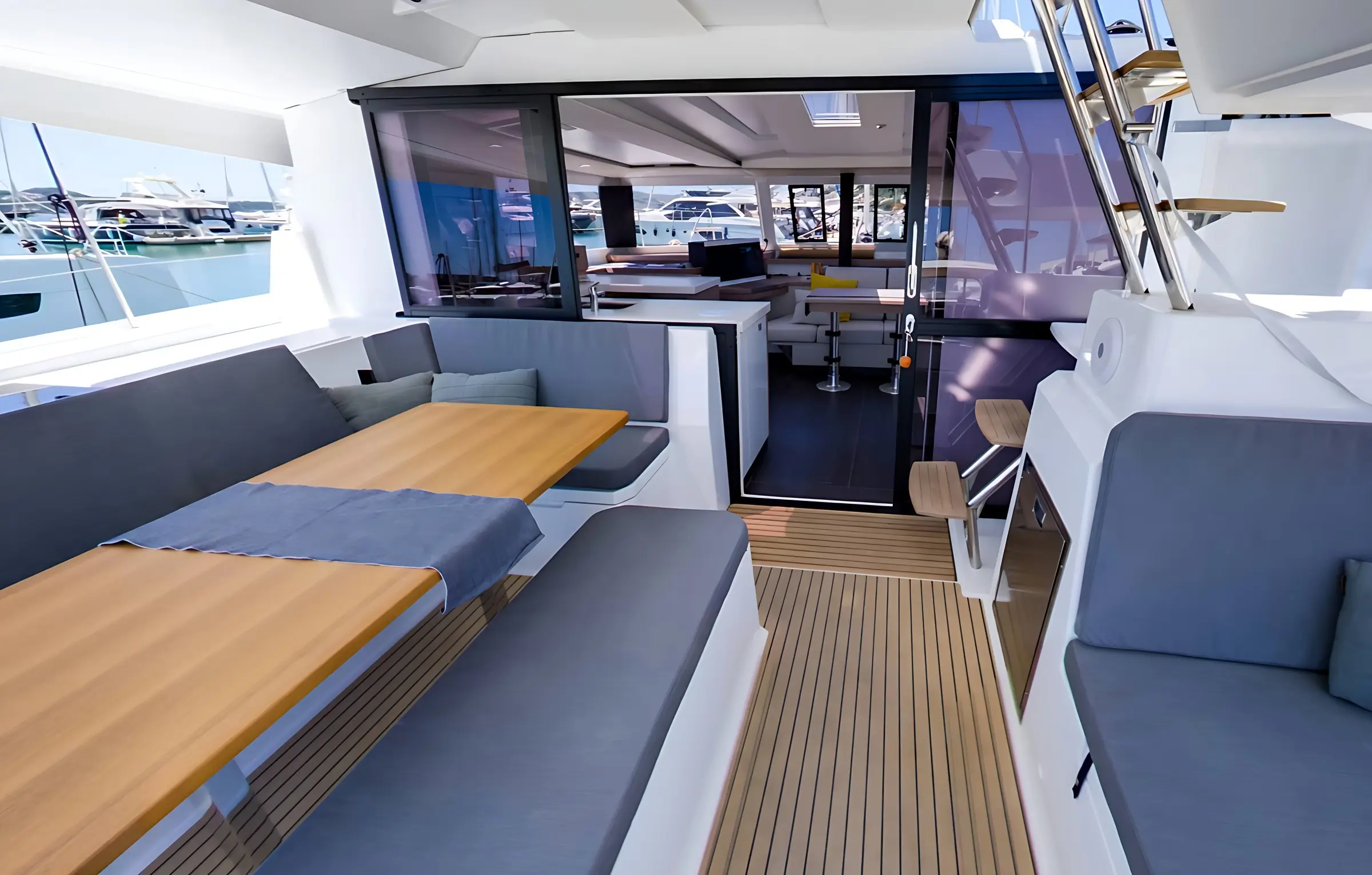 Modern yacht interior of the Fountaine Pajot Astrea 42 with cushioned seating, wooden tables, and open sliding doors leading to a spacious cabin with additional seating and a helm, docked at a marina with other boats visible outside.
