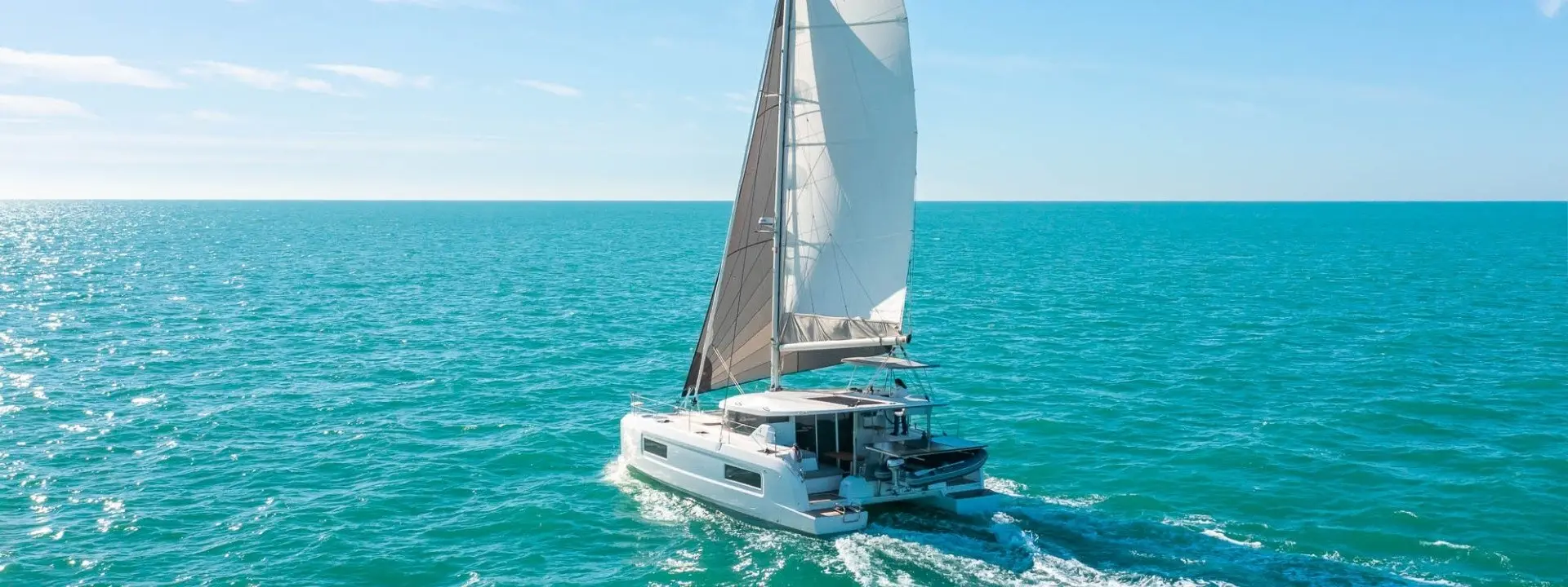 A white Lagoon 38 catamaran sailboat with tall sails glides on calm turquoise water under a clear blue sky, leaving a gentle wake behind.