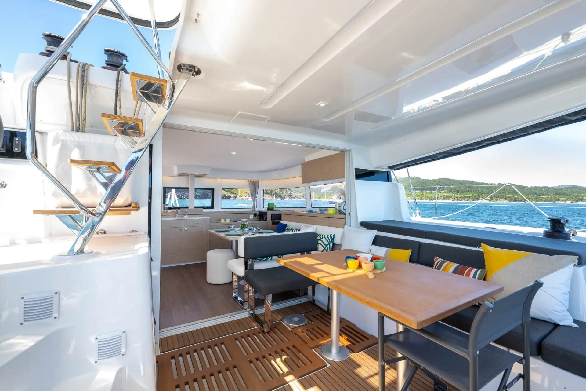 The interior of a modern yacht with a wooden table, cushioned seating, colorful pillows, and open access to a kitchen area; large windows offer a view of the sea and coastline.