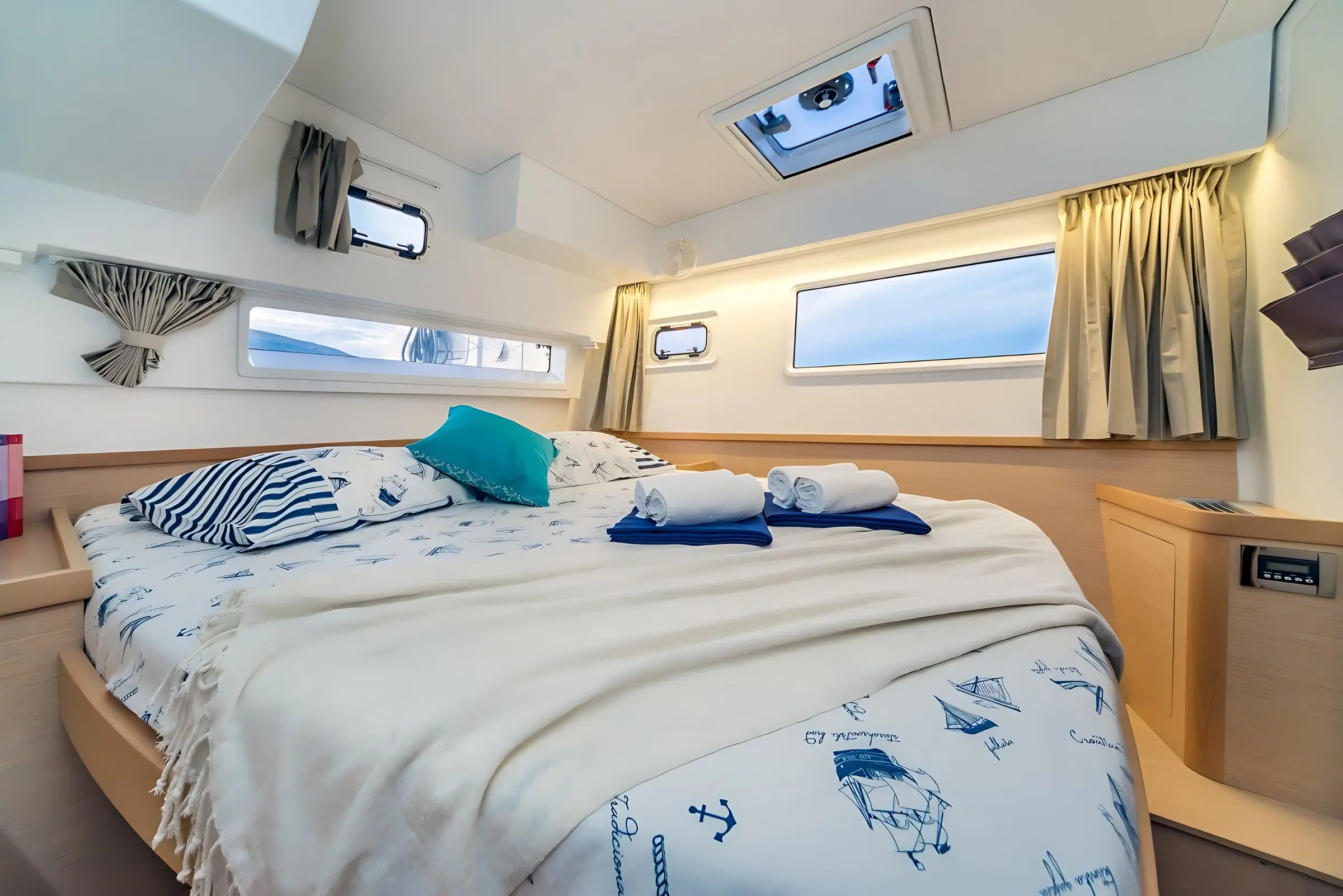 A cozy boat cabin bedroom with a double bed covered in nautical-themed bedding, two rolled towels, a blue pillow, light wood furniture, and several windows letting in natural light.