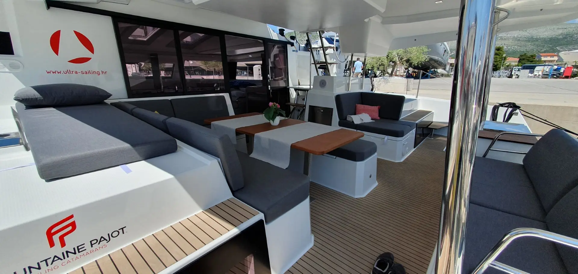 The image shows the spacious outdoor seating area of a modern Fountaine Pajot Elba 45 catamaran yacht, featuring gray cushioned benches, wooden tables, and decorative flowers, docked at a marina with land visible in the background.