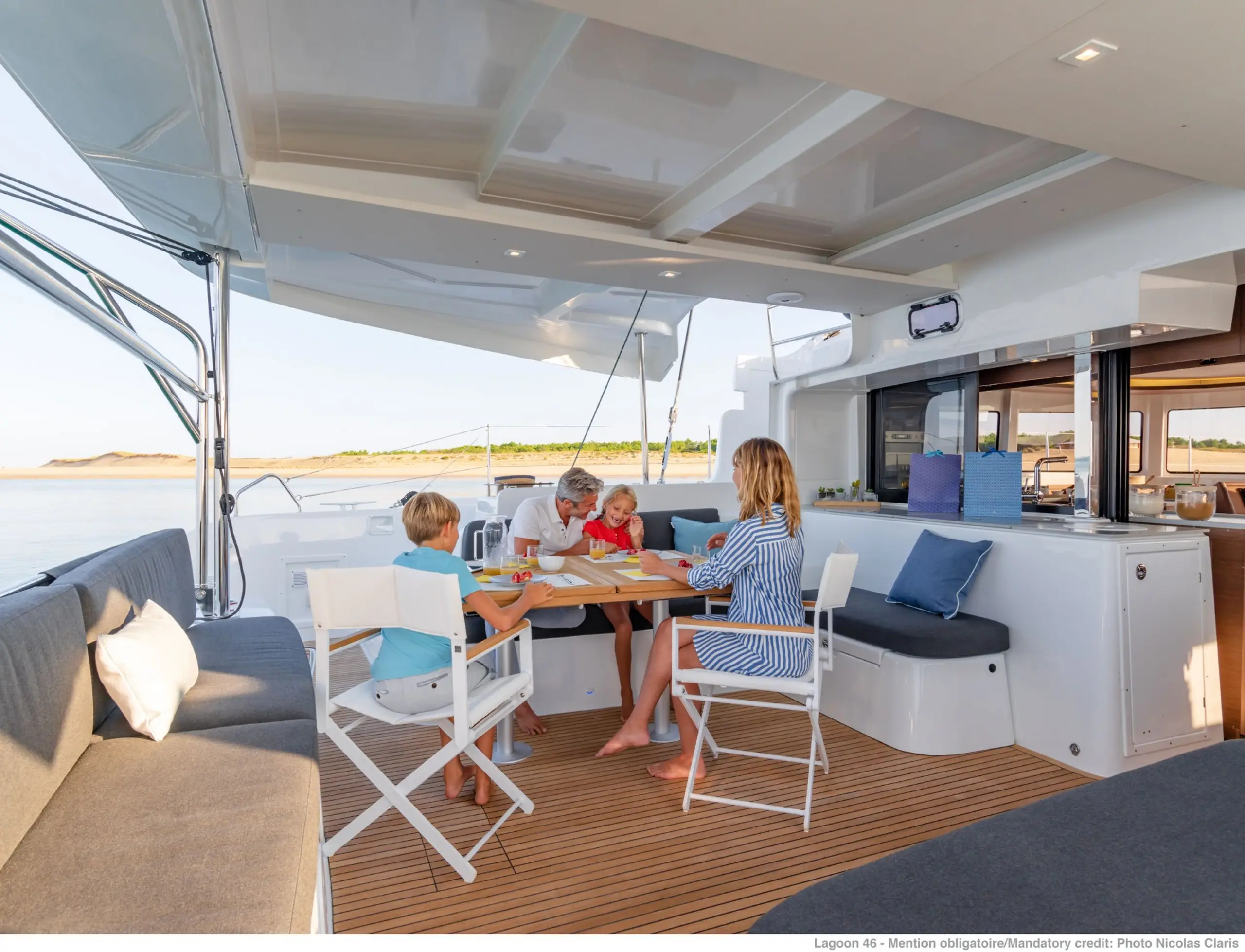 A family of four sits around a table on the deck of a modern Lagoon 46 yacht, enjoying breakfast near the water. The scene is bright and peaceful, with blue skies and a view of sandy shoreline in the background.