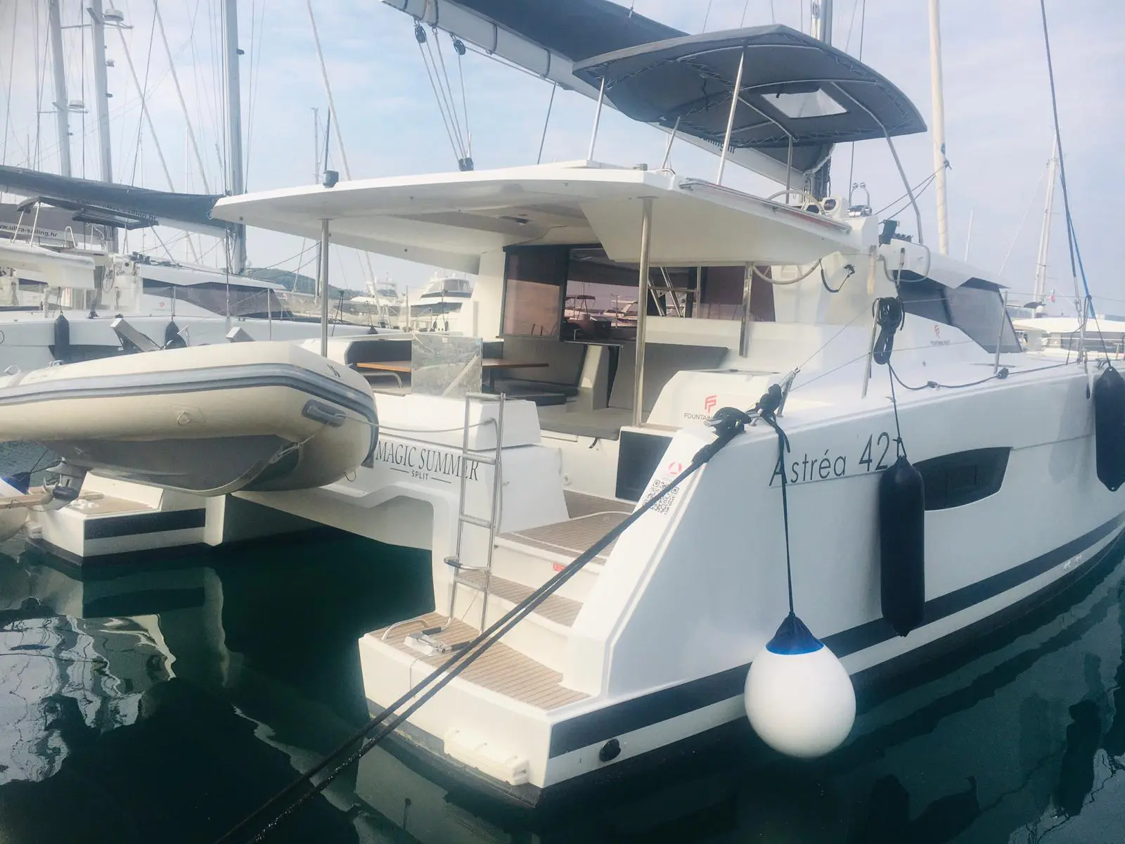 A white Fountaine Pajot Astrea 42 catamaran is docked at a marina, with an inflatable dinghy attached to the stern and black fenders hanging on the side. The deck and seating area are visible under a canopy.