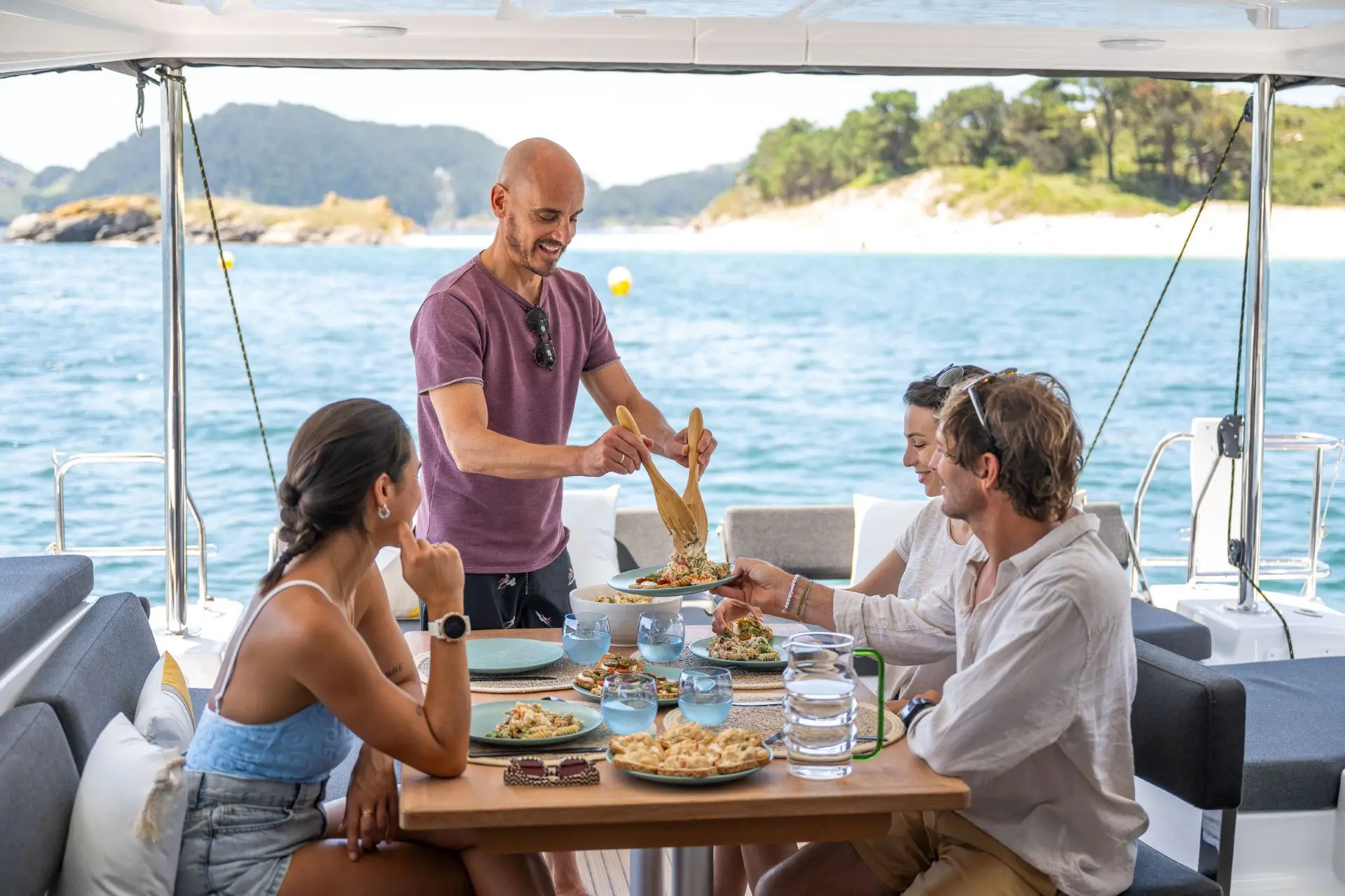 Four people sit around a table on a boat, enjoying food and drinks. One man serves a dish while the others smile. The boat is near a scenic coast with trees and clear blue water in the background.