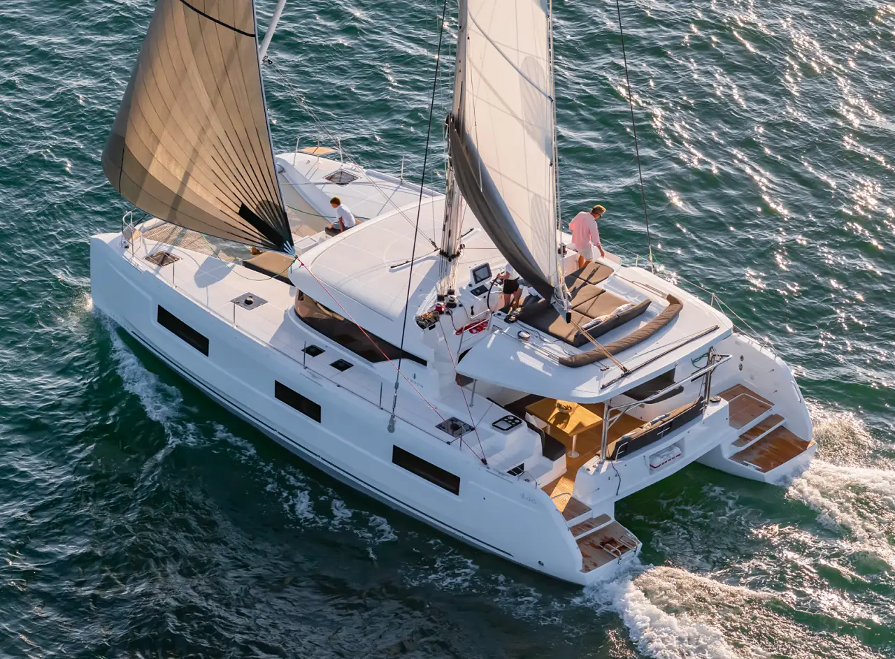 A white Lagoon 46 catamaran sailboat with two people onboard sails on clear, sunlit water. The sails are raised, and the boat leaves a small wake behind it as it moves forward.