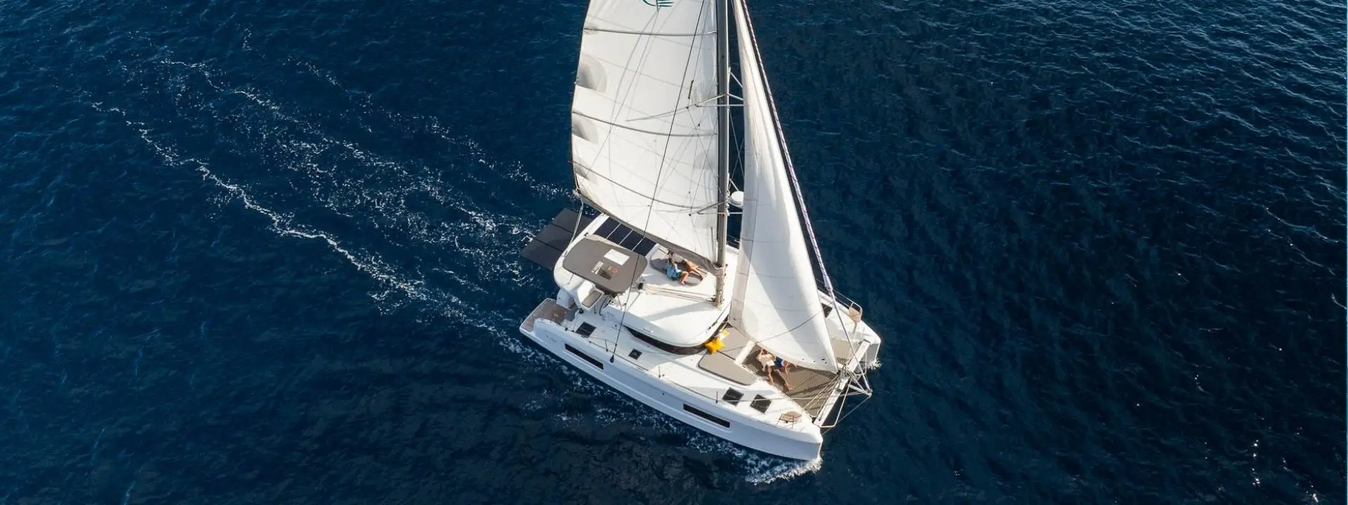 Aerial view of a Lagoon 38 white sailboat with its sails up, cutting through deep blue ocean water, creating small waves as it moves forward.