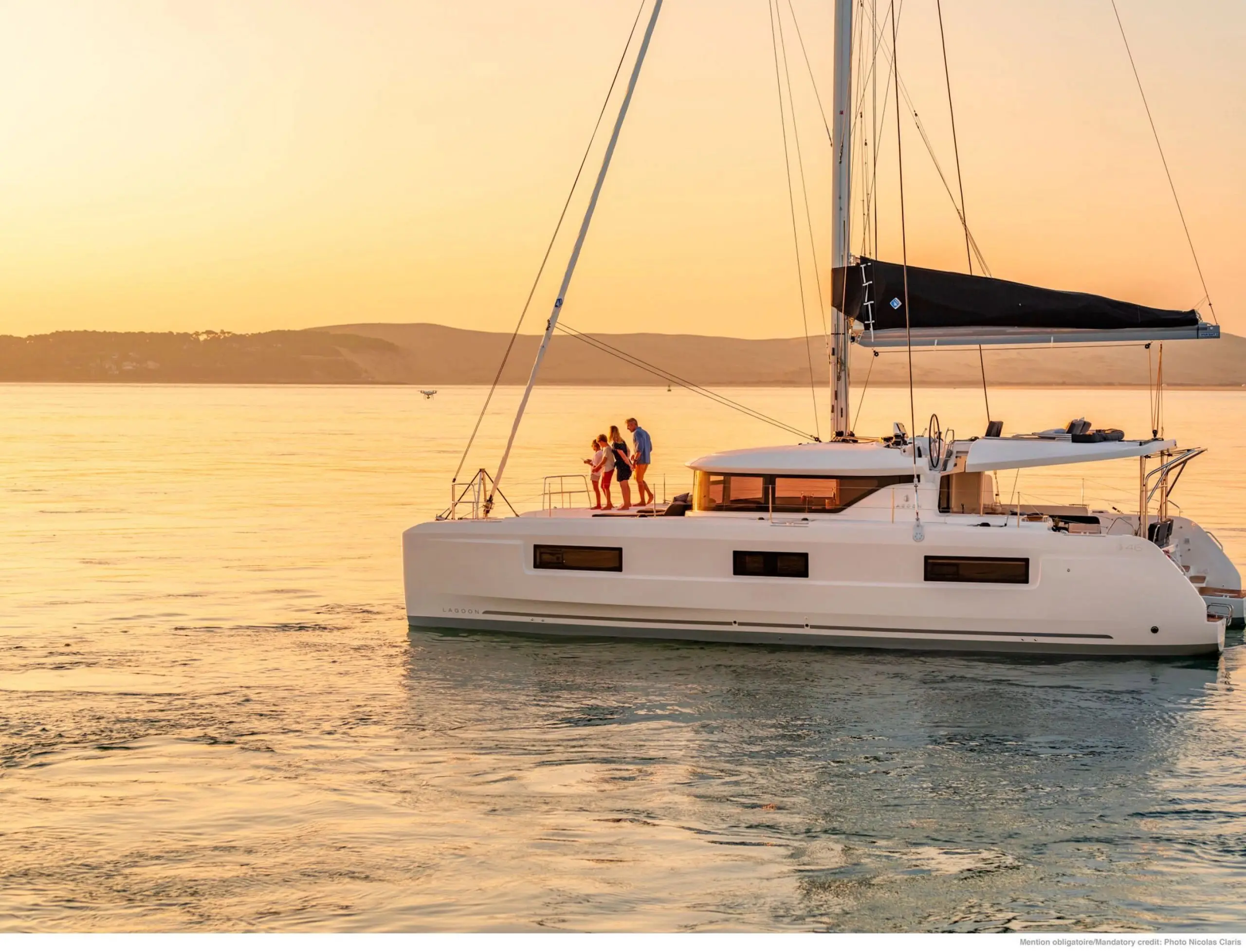 A family stands together on the deck of a white Lagoon 46 sailboat at sunset, enjoying the calm water and golden sky. Rolling hills are visible in the distance.