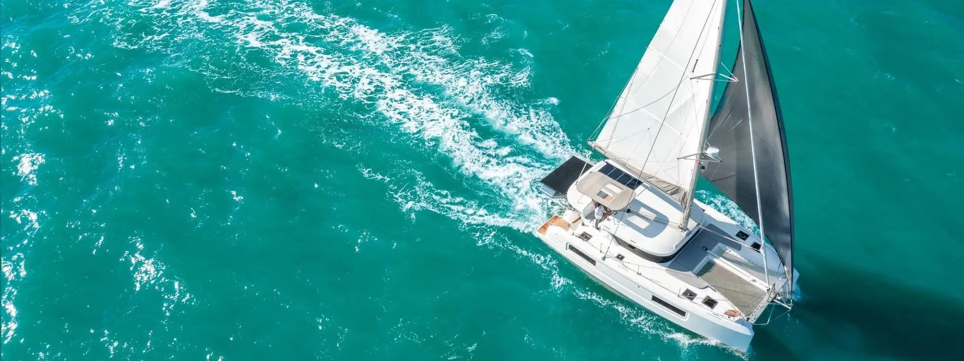 Aerial view of a Lagoon 38 white sailboat sailing through clear turquoise water, leaving a foamy trail behind as it moves forward under bright sunlight.