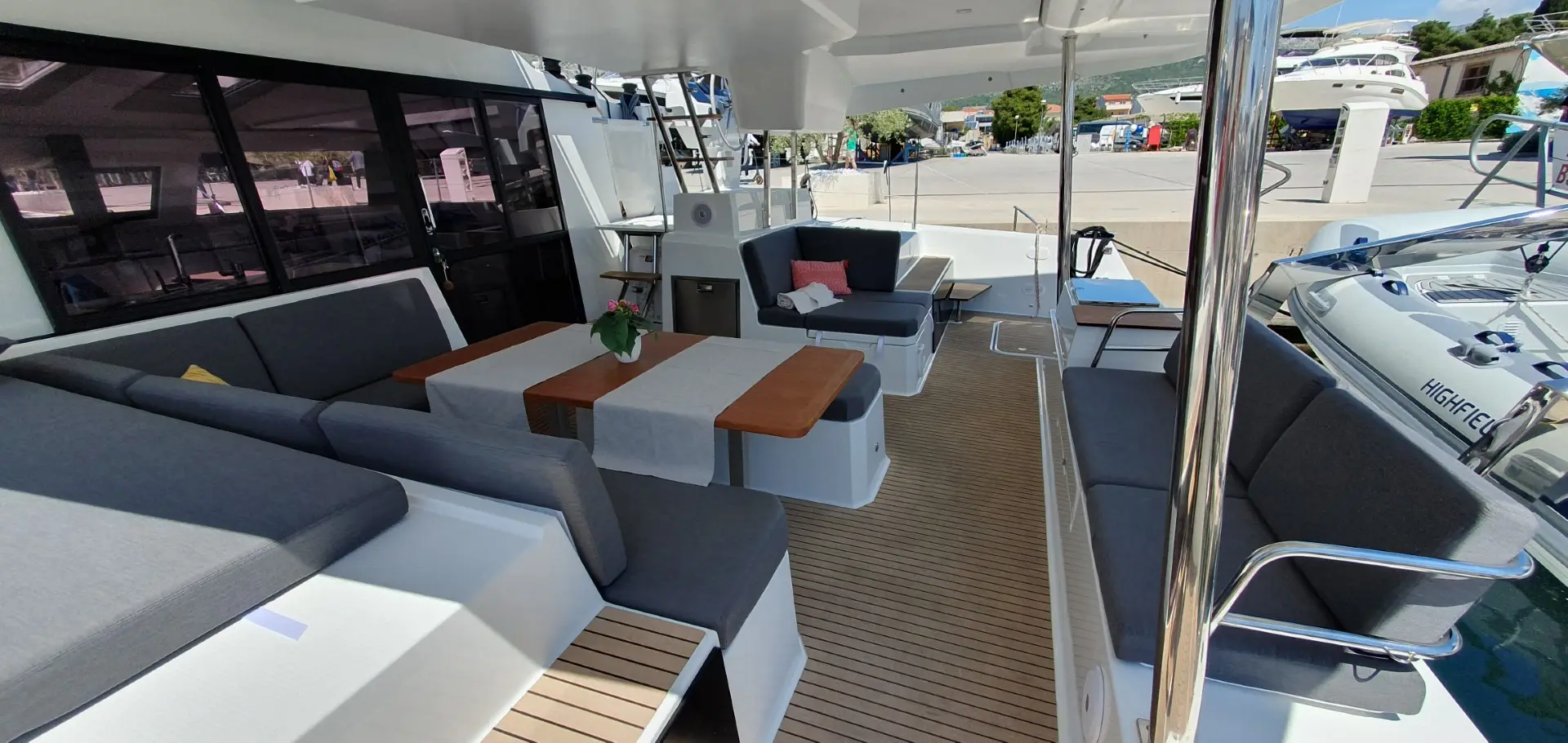 Spacious outdoor seating area on a Fountaine Pajot Elba 45 yacht with gray cushioned benches, wooden dining table, potted flowers, and decorative pillows. The yacht is docked at a marina with boats and buildings visible in the background.