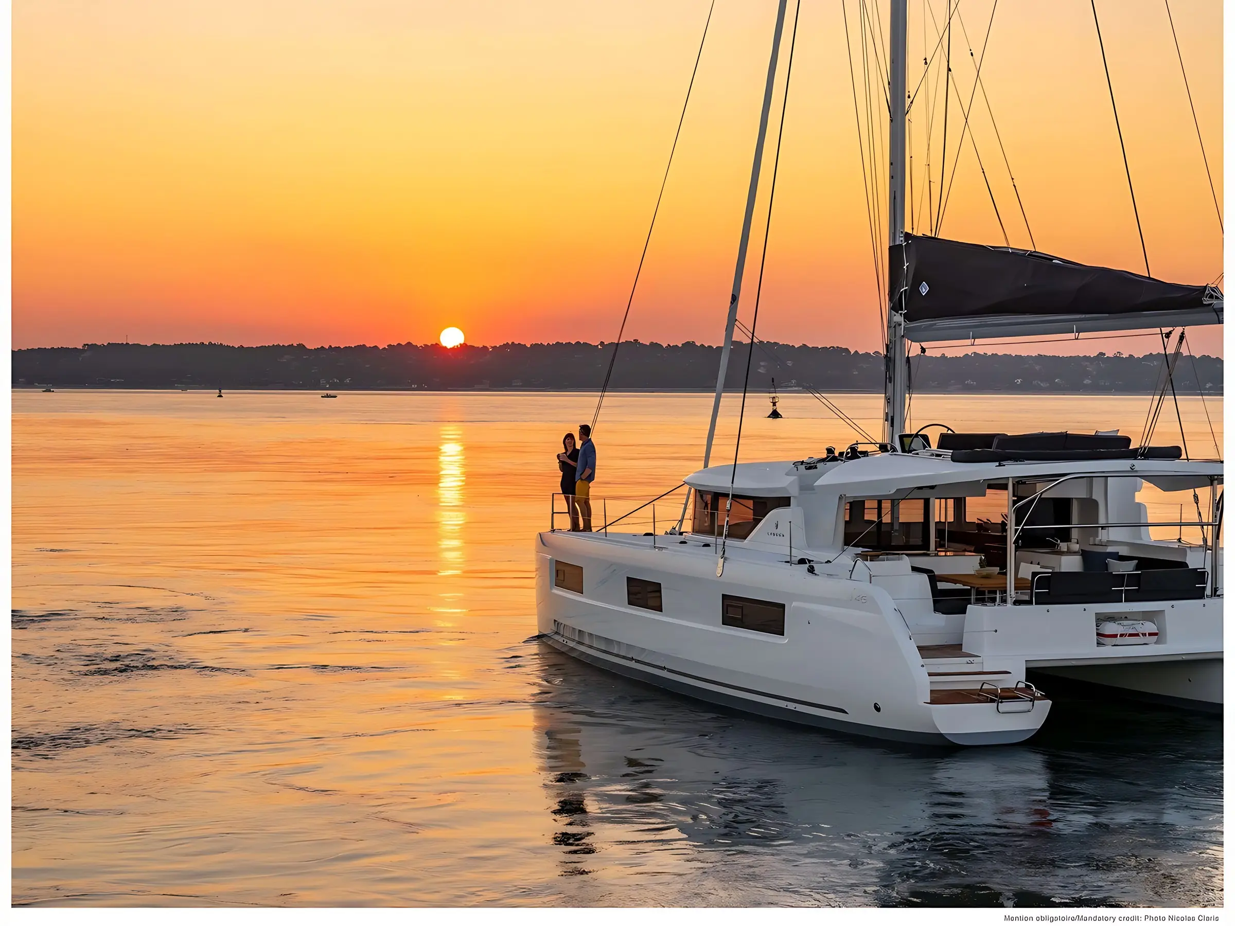 A couple stands on the deck of a white Lagoon 46 catamaran yacht at sunset, with calm water and an orange sky in the background. The sun sets near the horizon, casting a warm glow over the serene scene.