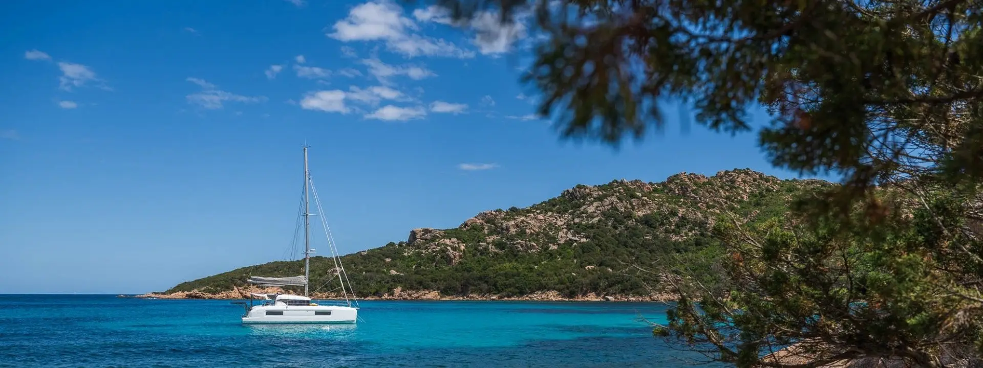 A white Lagoon 38 sailboat floats on clear turquoise water near a rocky, green hillside under a blue sky, framed by tree branches in the foreground.