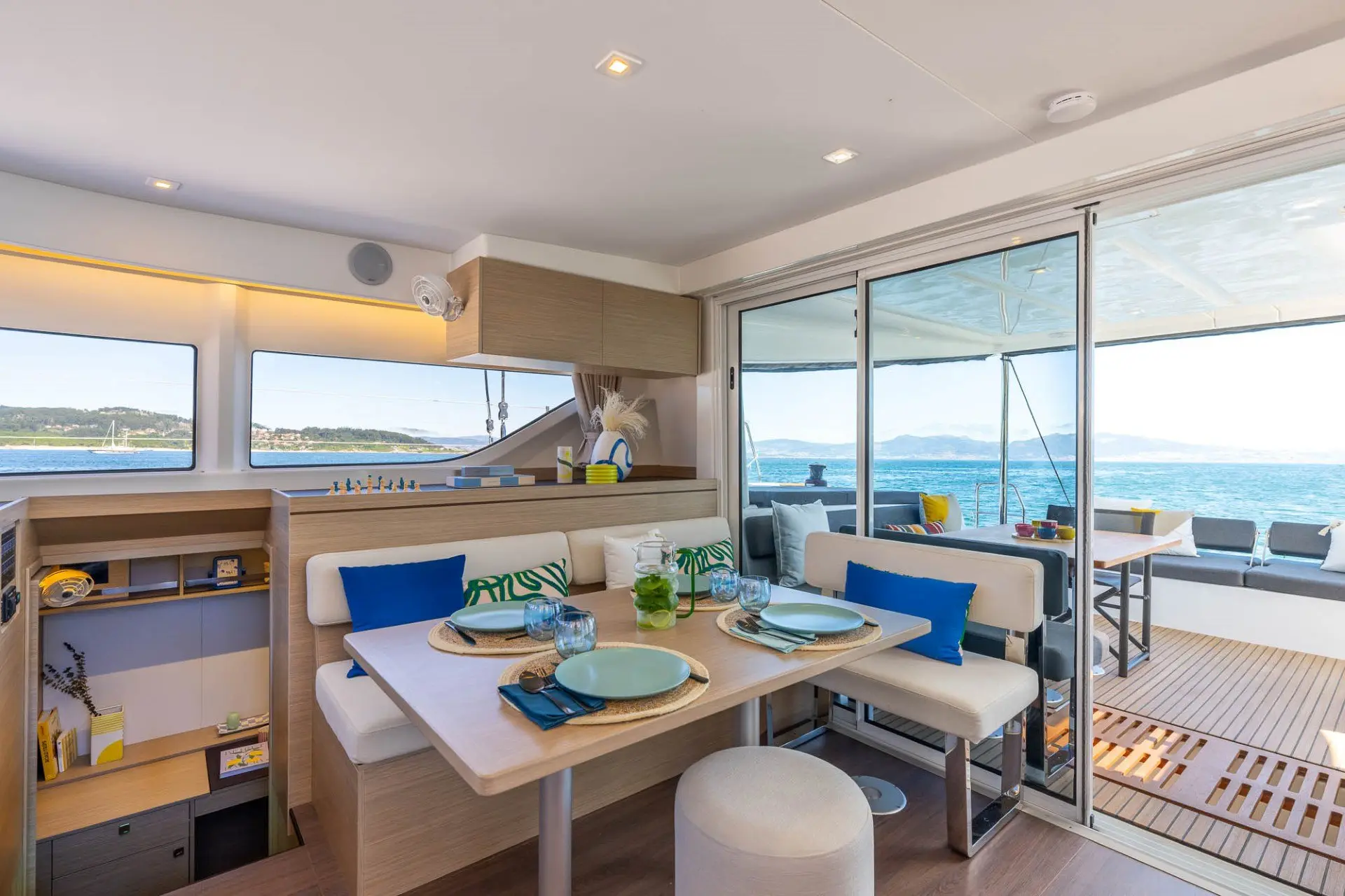 The interior of a modern yacht with a dining table set for four, cushioned seating, large windows, and sliding glass doors opening to a deck with ocean views.