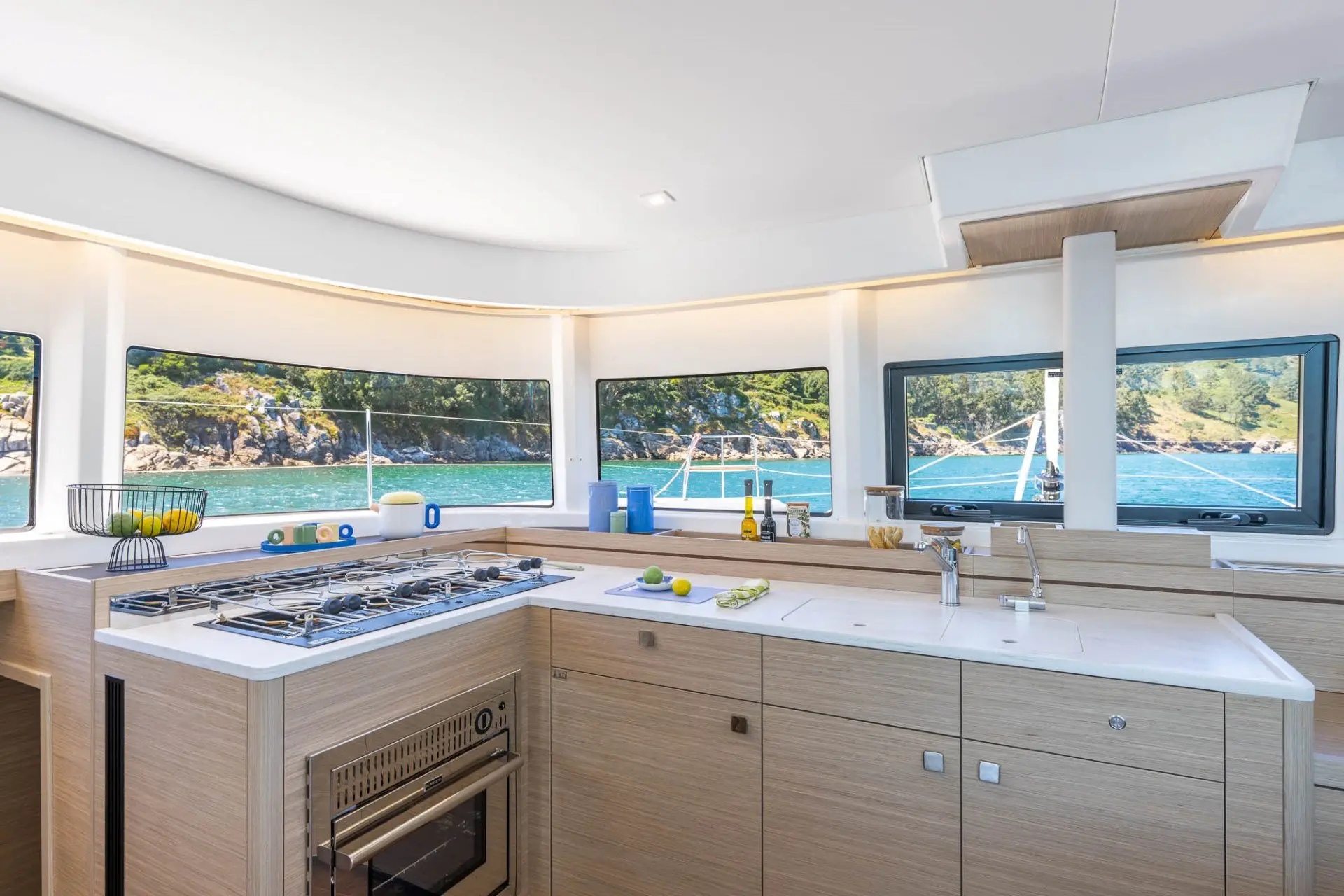 Modern yacht kitchen with light wood cabinetry, a stove, oven, sink, and countertops. Windows offer views of turquoise water and green shoreline outside, with kitchen items and fresh ingredients arranged neatly inside.